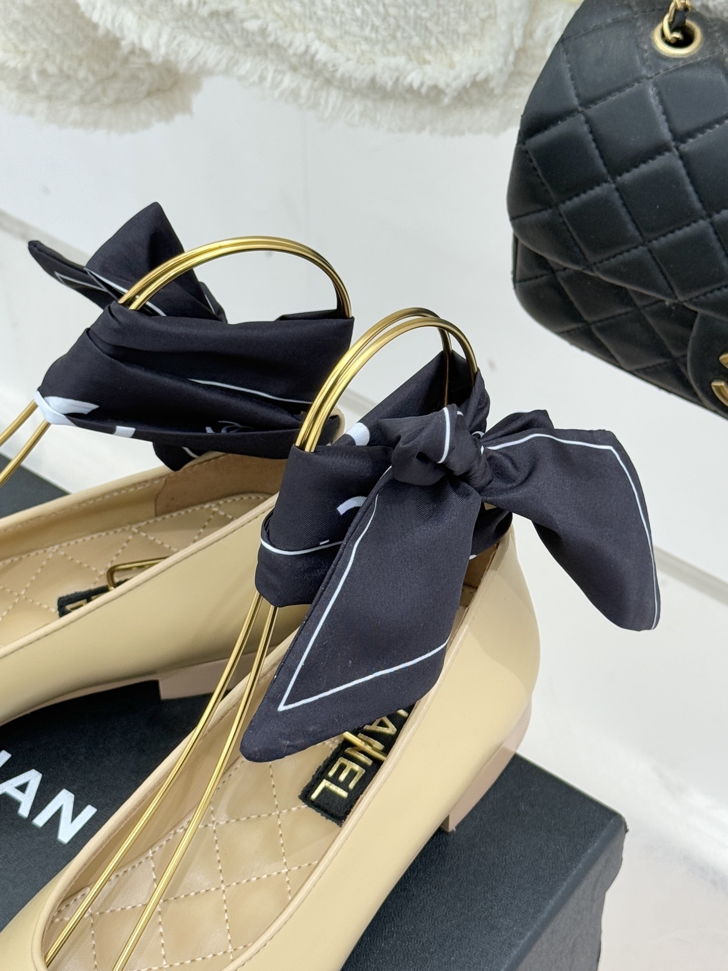 A pair of Chanel beige lambskin ballerina flats with black cap toes and decorative silk bows placed on a black shoe box. - Hình 8