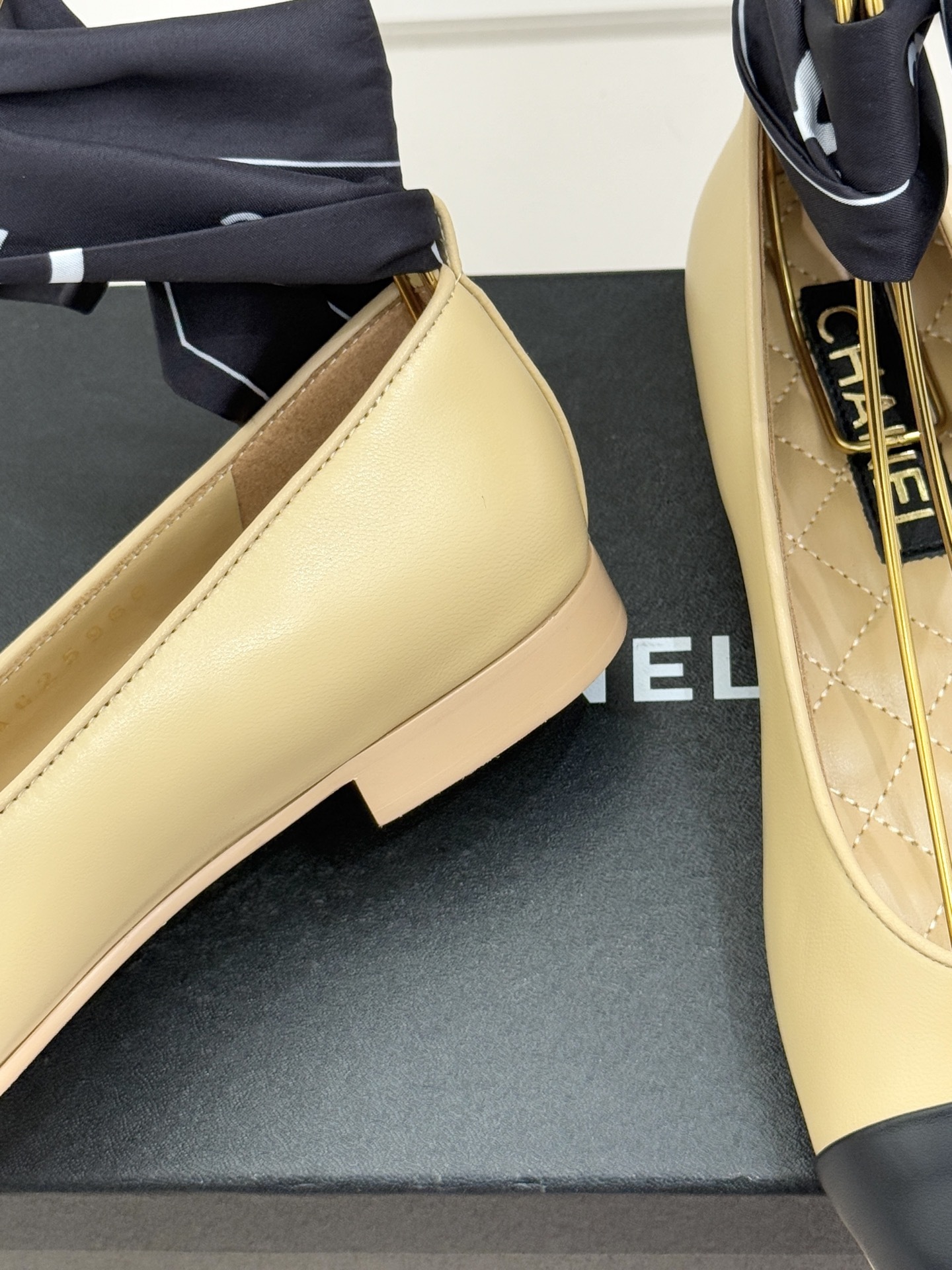 A pair of Chanel beige lambskin ballerina flats with black cap toes and decorative silk bows placed on a black shoe box. - Hình 6