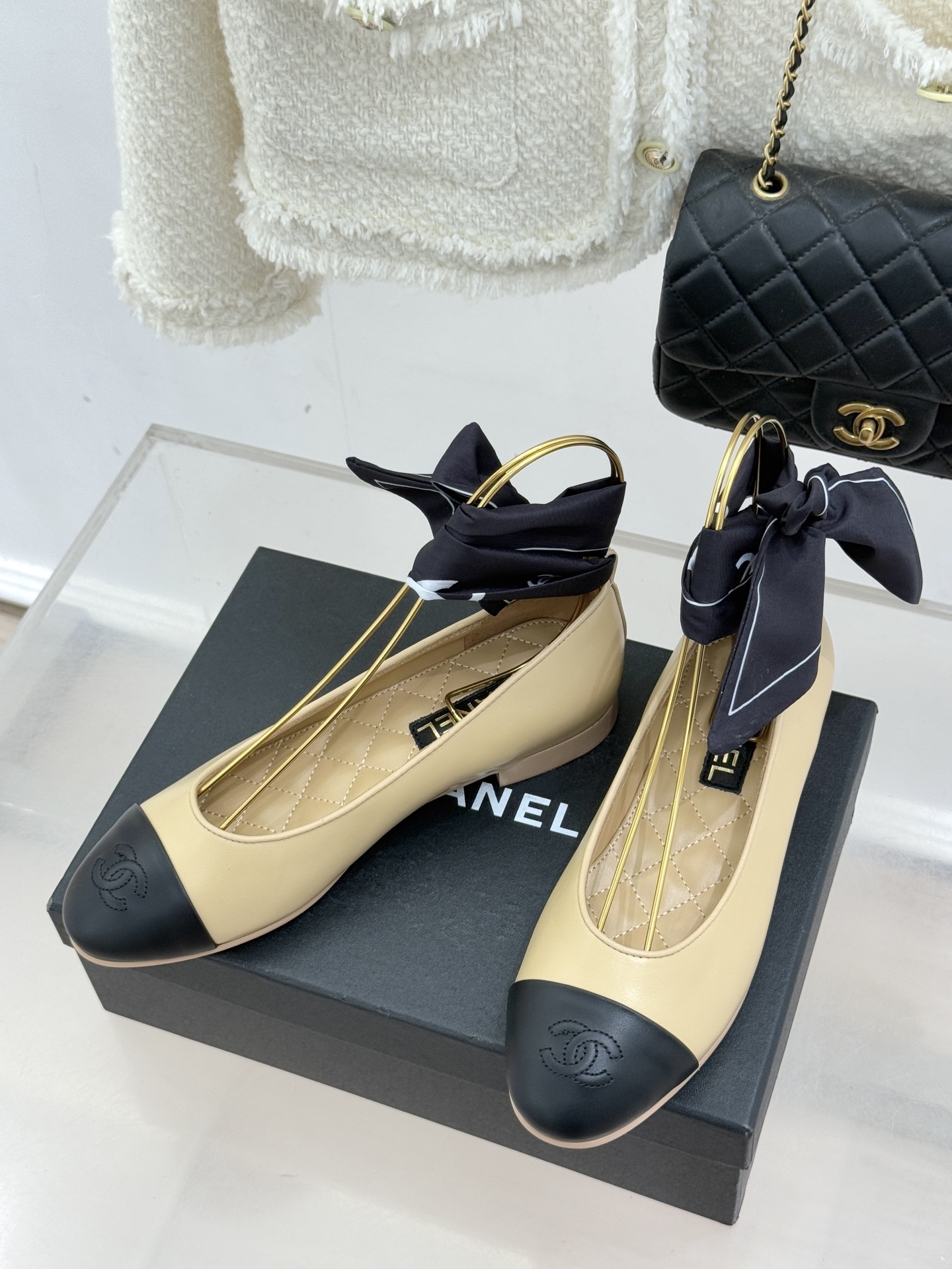 A pair of Chanel beige lambskin ballerina flats with black cap toes and decorative silk bows placed on a black shoe box. - Hình 2