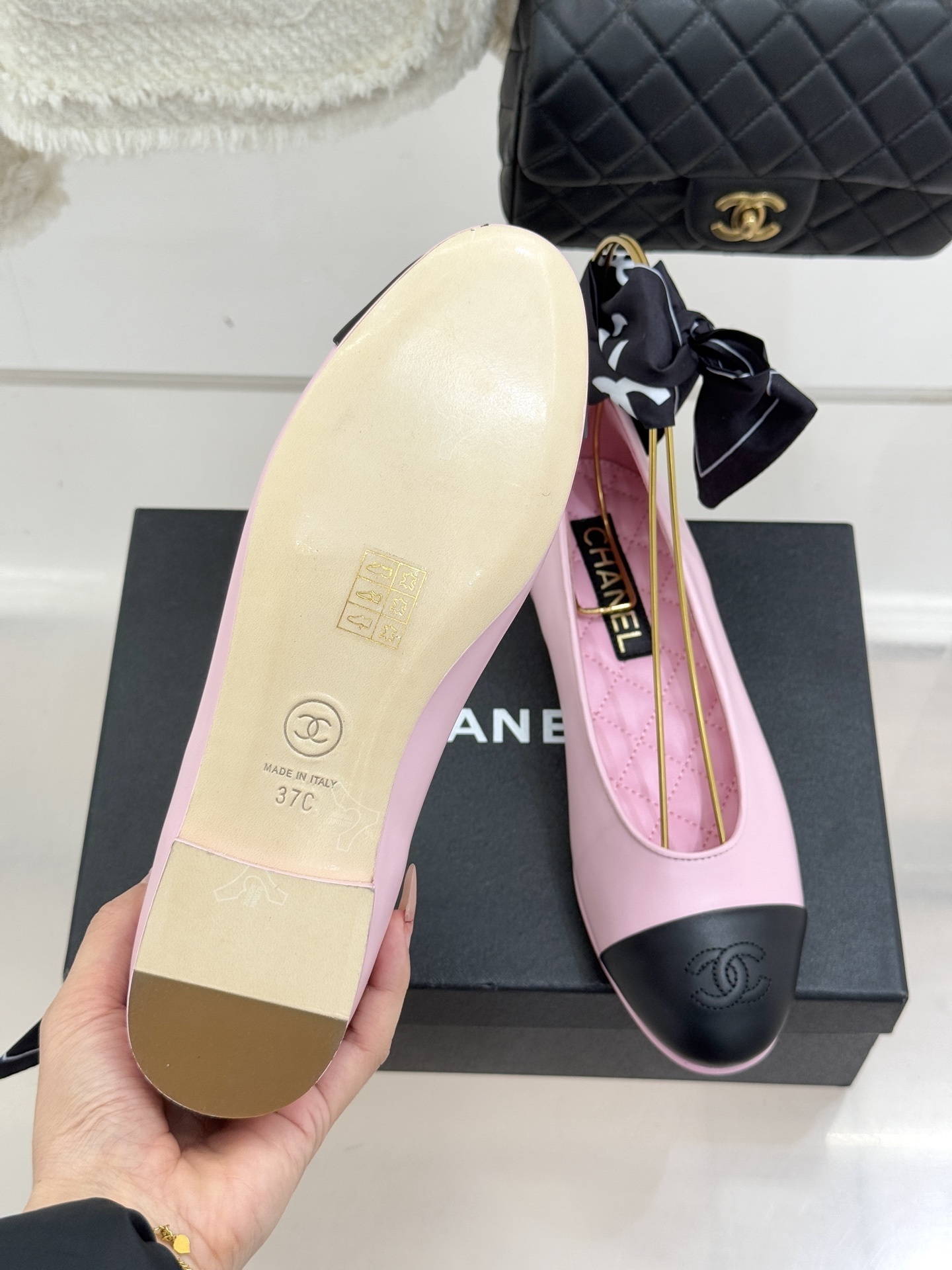 A pair of pink Chanel ballet flats with black leather cap toes featuring the CC logo, resting on a black shoe box next to a tweed jacket and quilted bag. - Hình 9