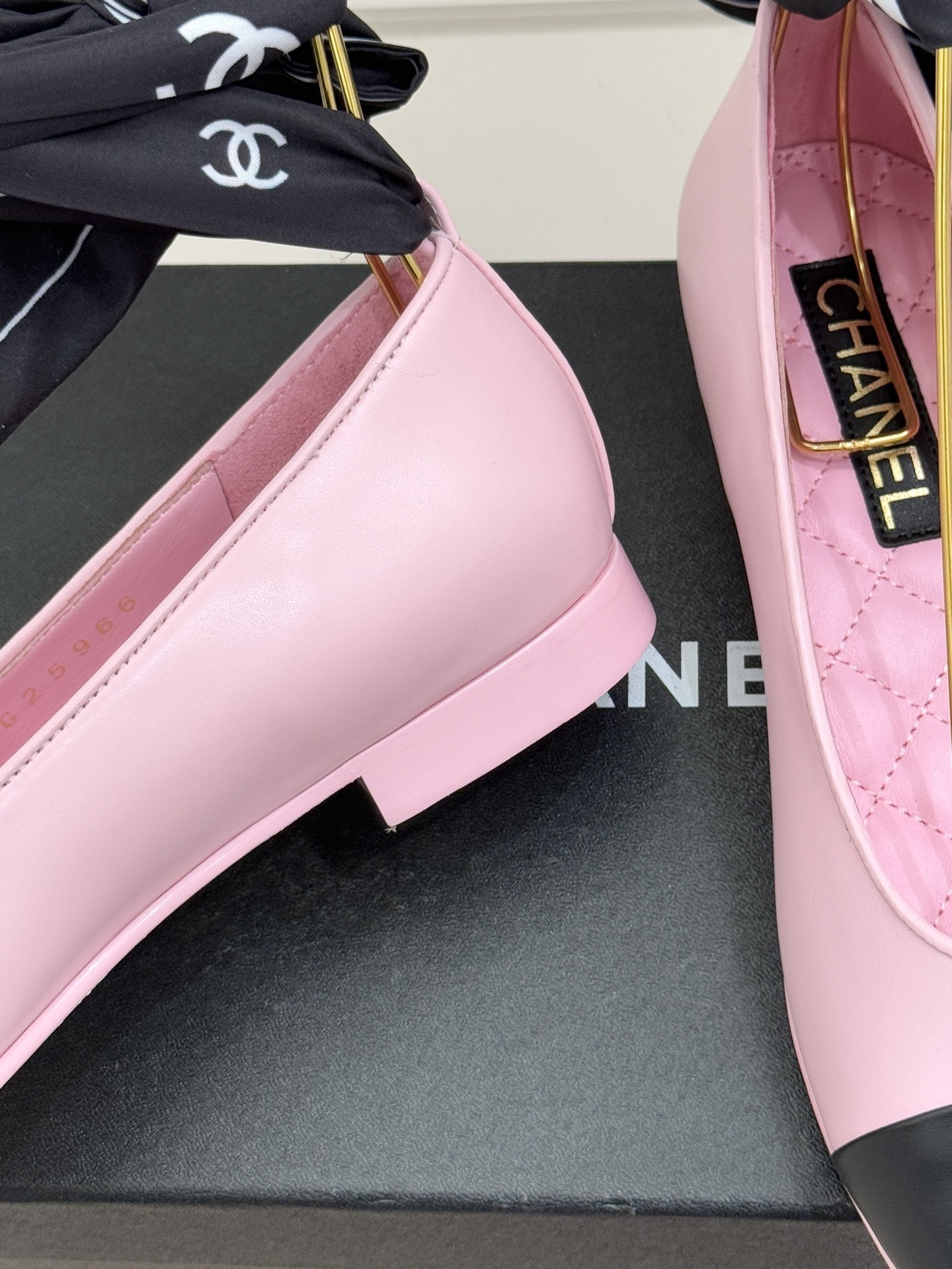 A pair of pink Chanel ballet flats with black leather cap toes featuring the CC logo, resting on a black shoe box next to a tweed jacket and quilted bag. - Hình 6
