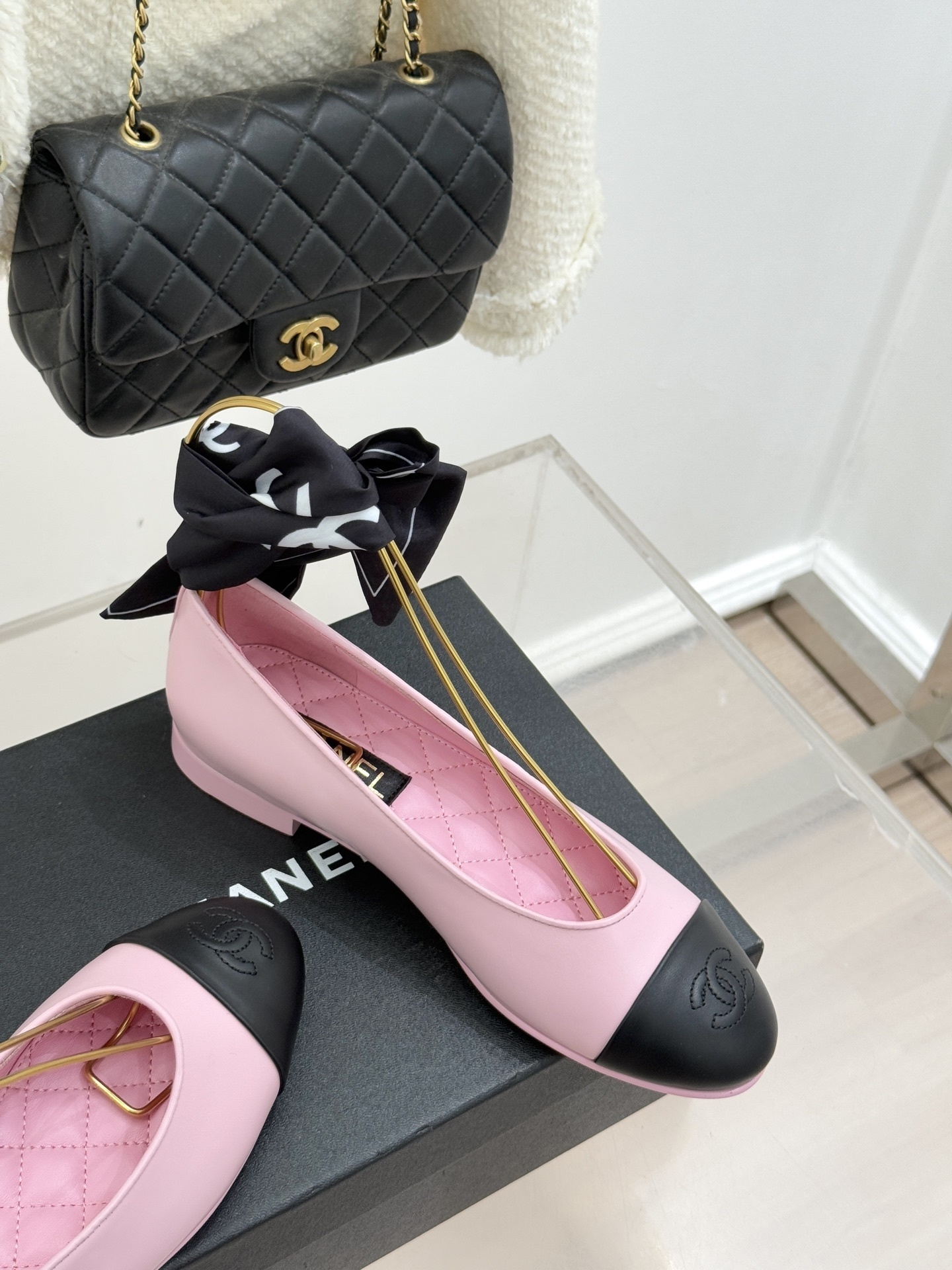 A pair of pink Chanel ballet flats with black leather cap toes featuring the CC logo, resting on a black shoe box next to a tweed jacket and quilted bag. - Hình 5