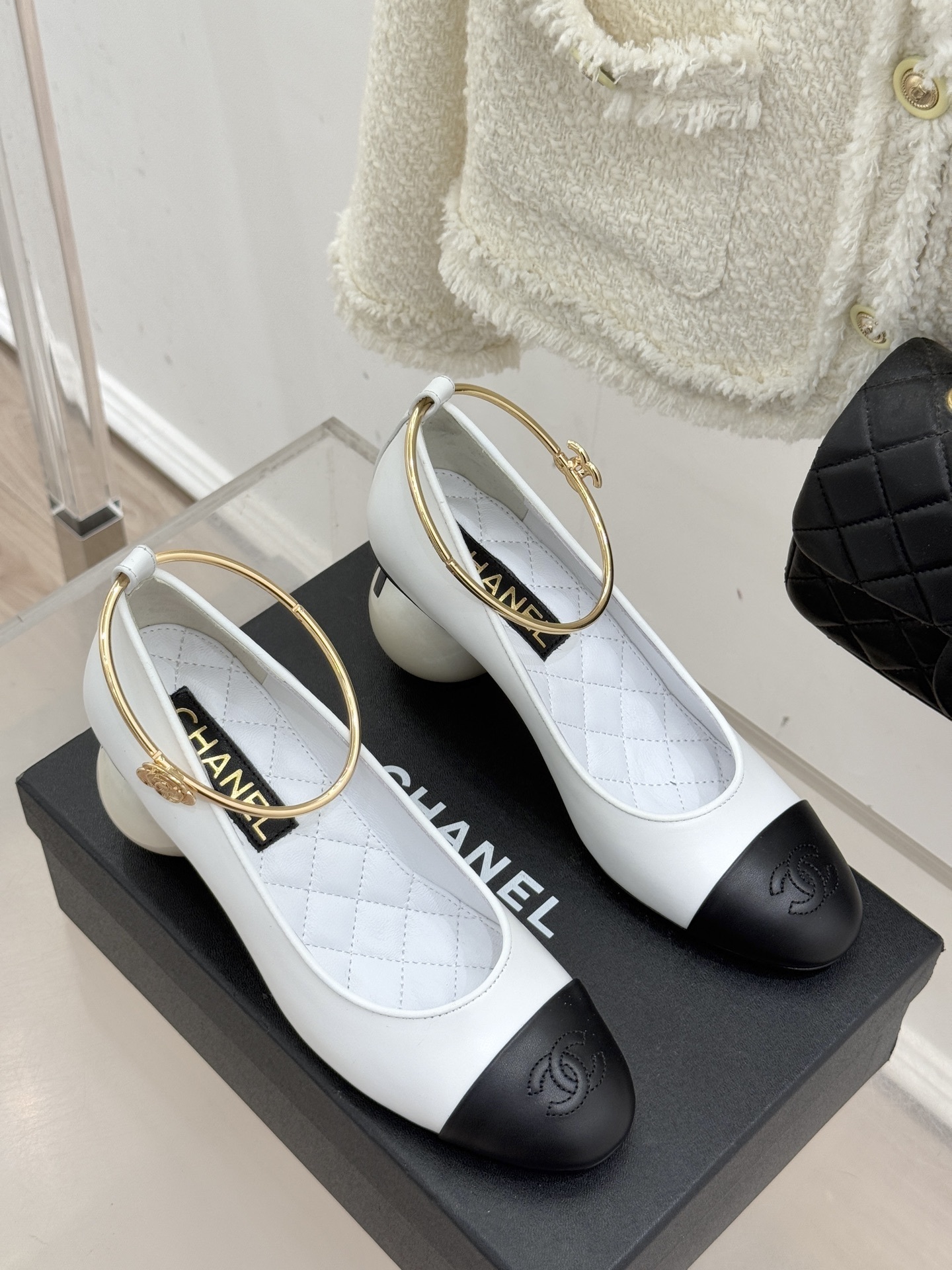Pair of Chanel white and black bicolor pumps with a spherical pearl heel and gold-tone ankle rings displayed on a black box. - Hình 3
