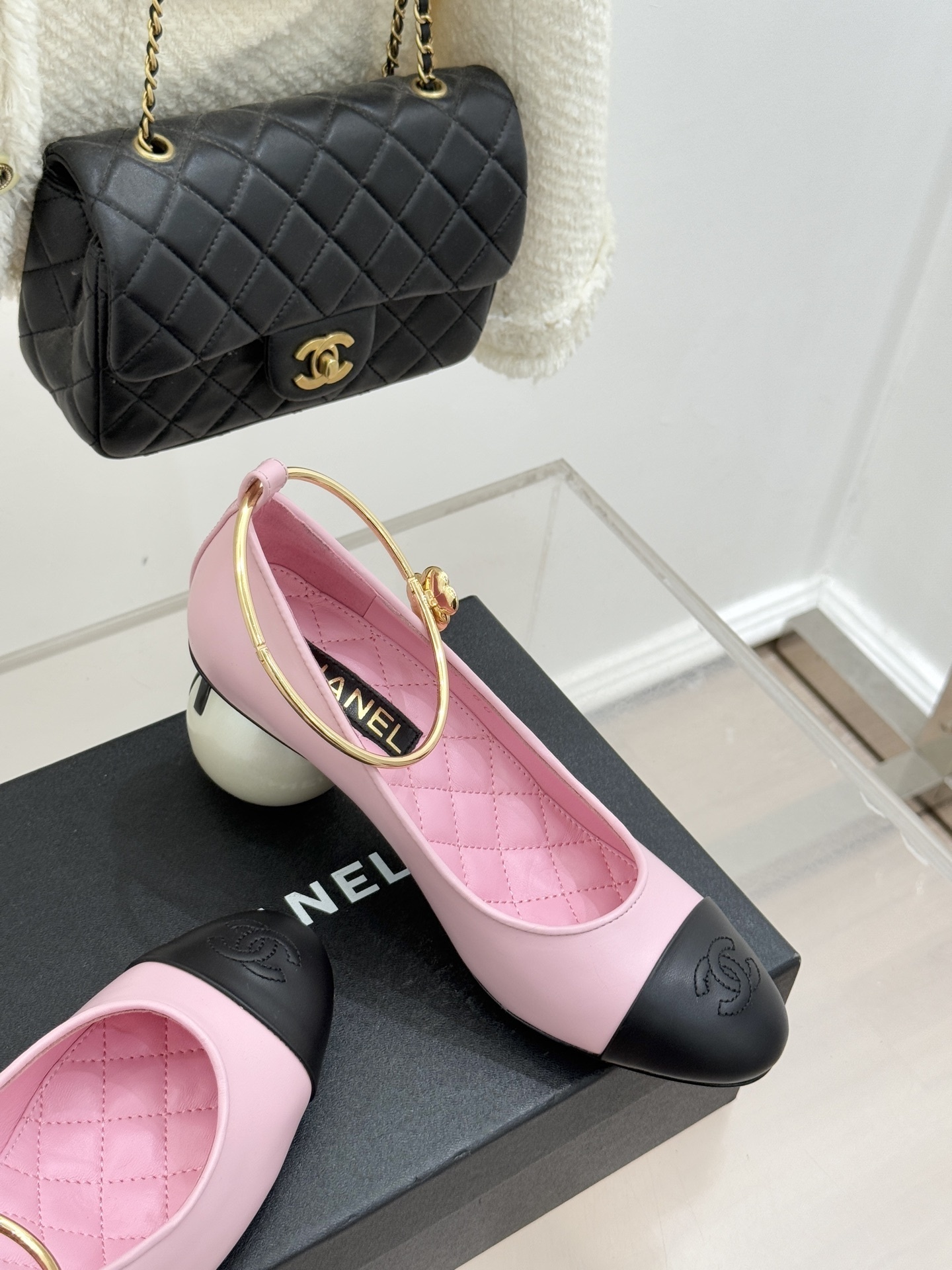 A pair of luxury pink Chanel pumps with black cap-toes, gold ankle ring details, and large pearl-shaped heels resting on a black branded box. - Hình 5