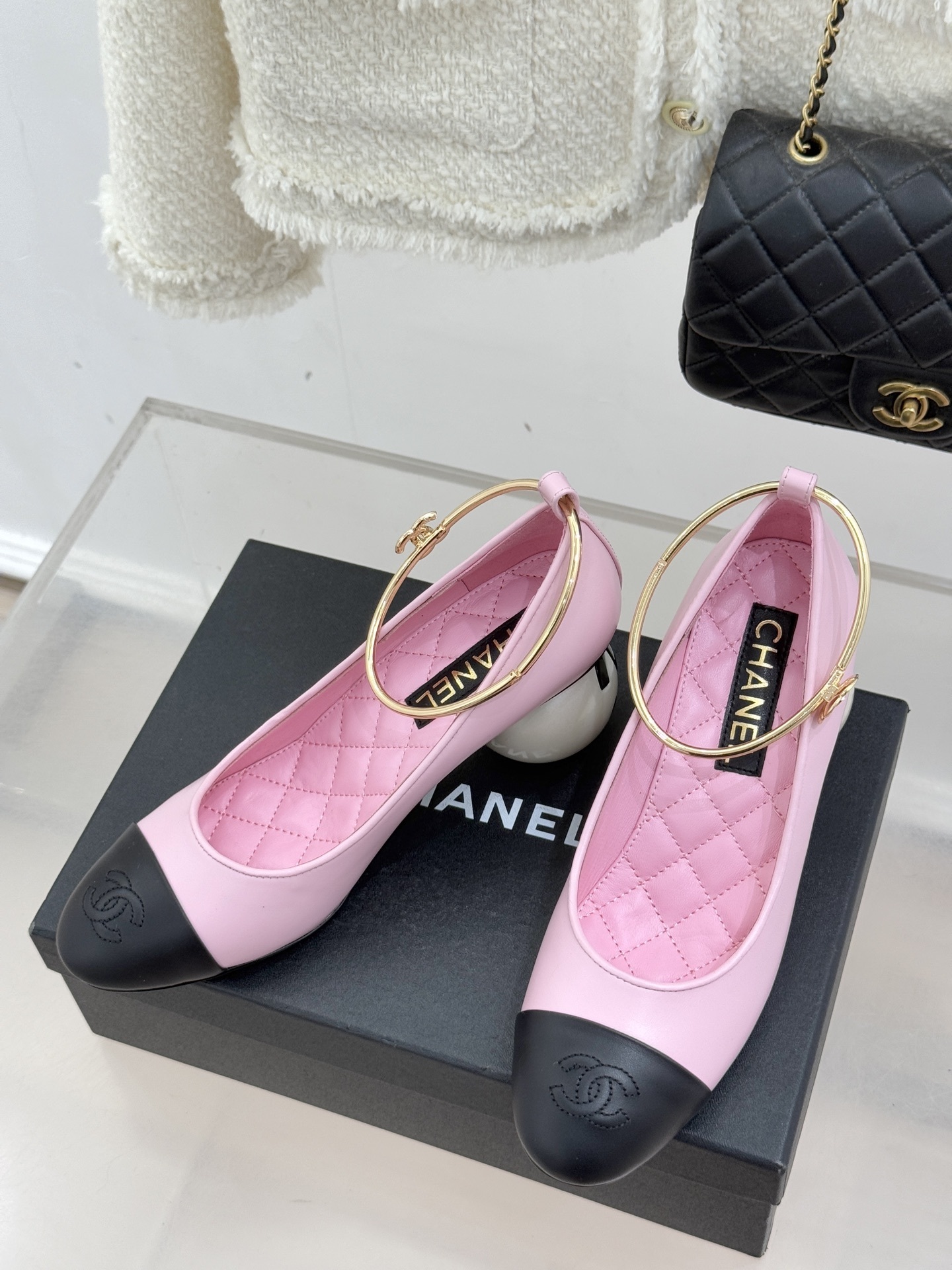A pair of luxury pink Chanel pumps with black cap-toes, gold ankle ring details, and large pearl-shaped heels resting on a black branded box. - Hình 2