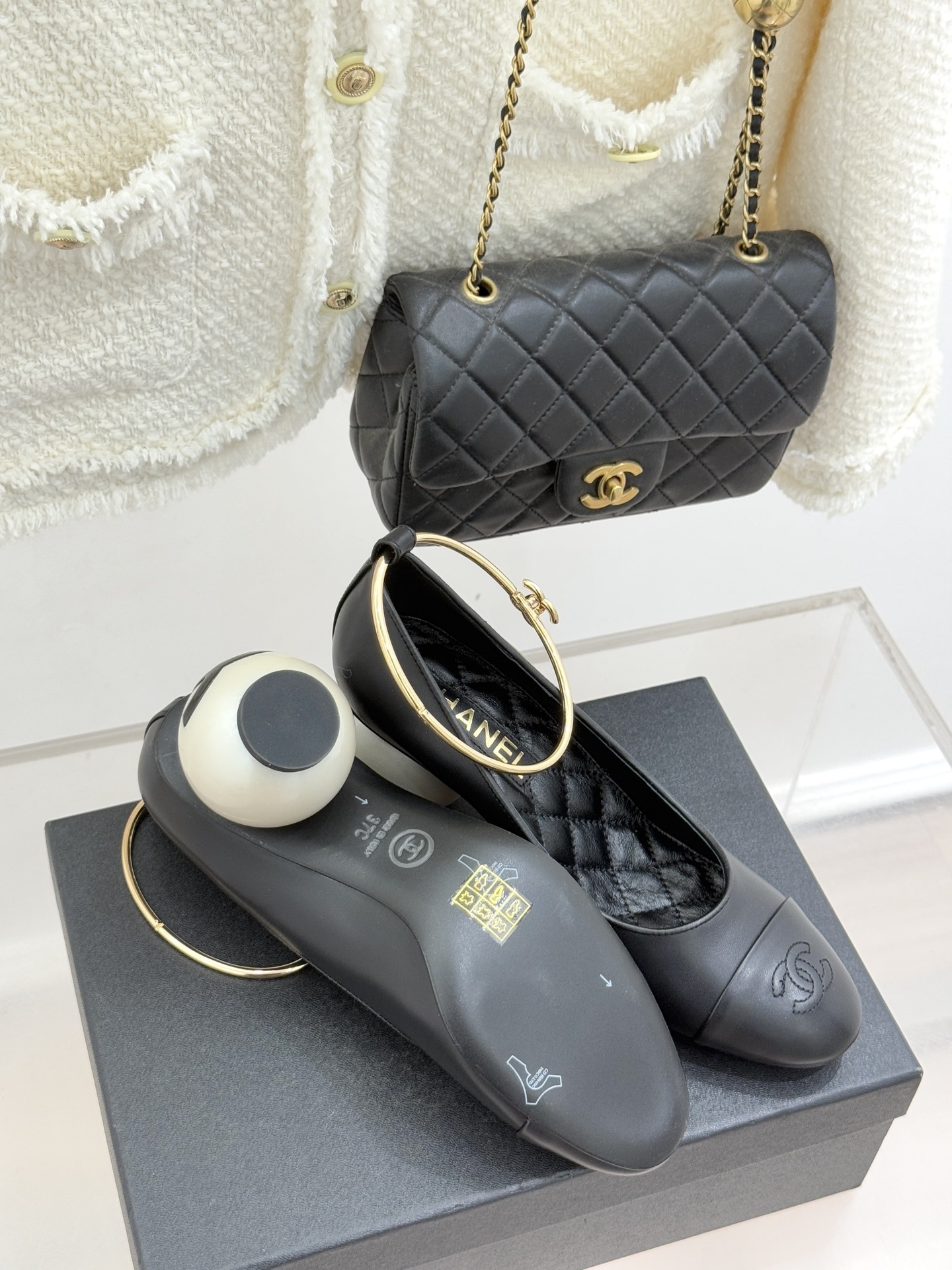 Pair of Chanel black leather pumps featuring a white round pearl heel and a gold-tone CC logo ankle ring on a black designer box. - Hình 9