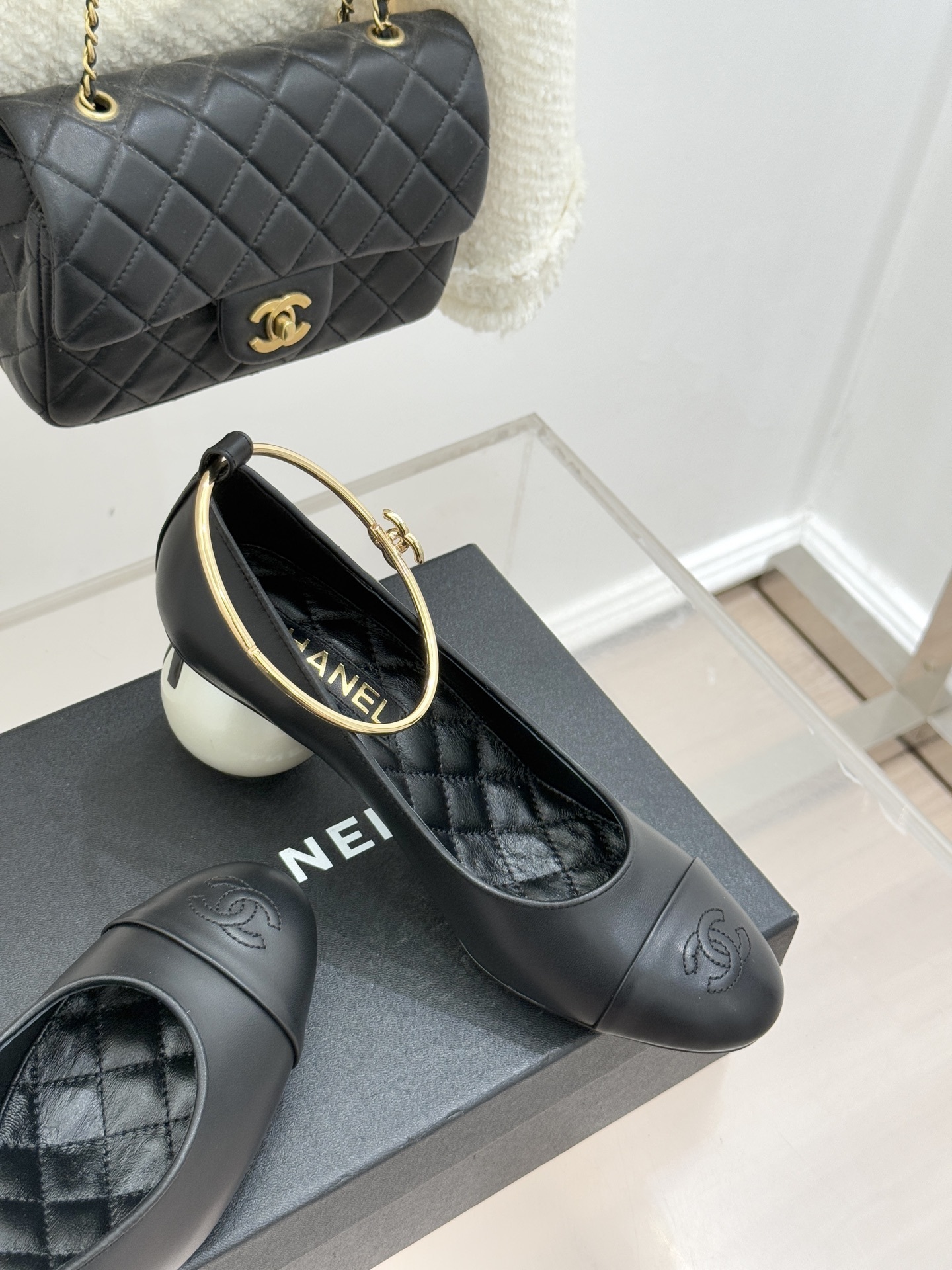 Pair of Chanel black leather pumps featuring a white round pearl heel and a gold-tone CC logo ankle ring on a black designer box. - Hình 5