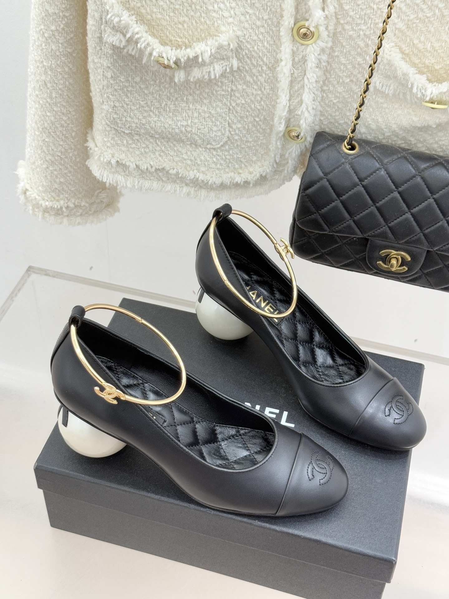 Pair of Chanel black leather pumps featuring a white round pearl heel and a gold-tone CC logo ankle ring on a black designer box. - Hình 3