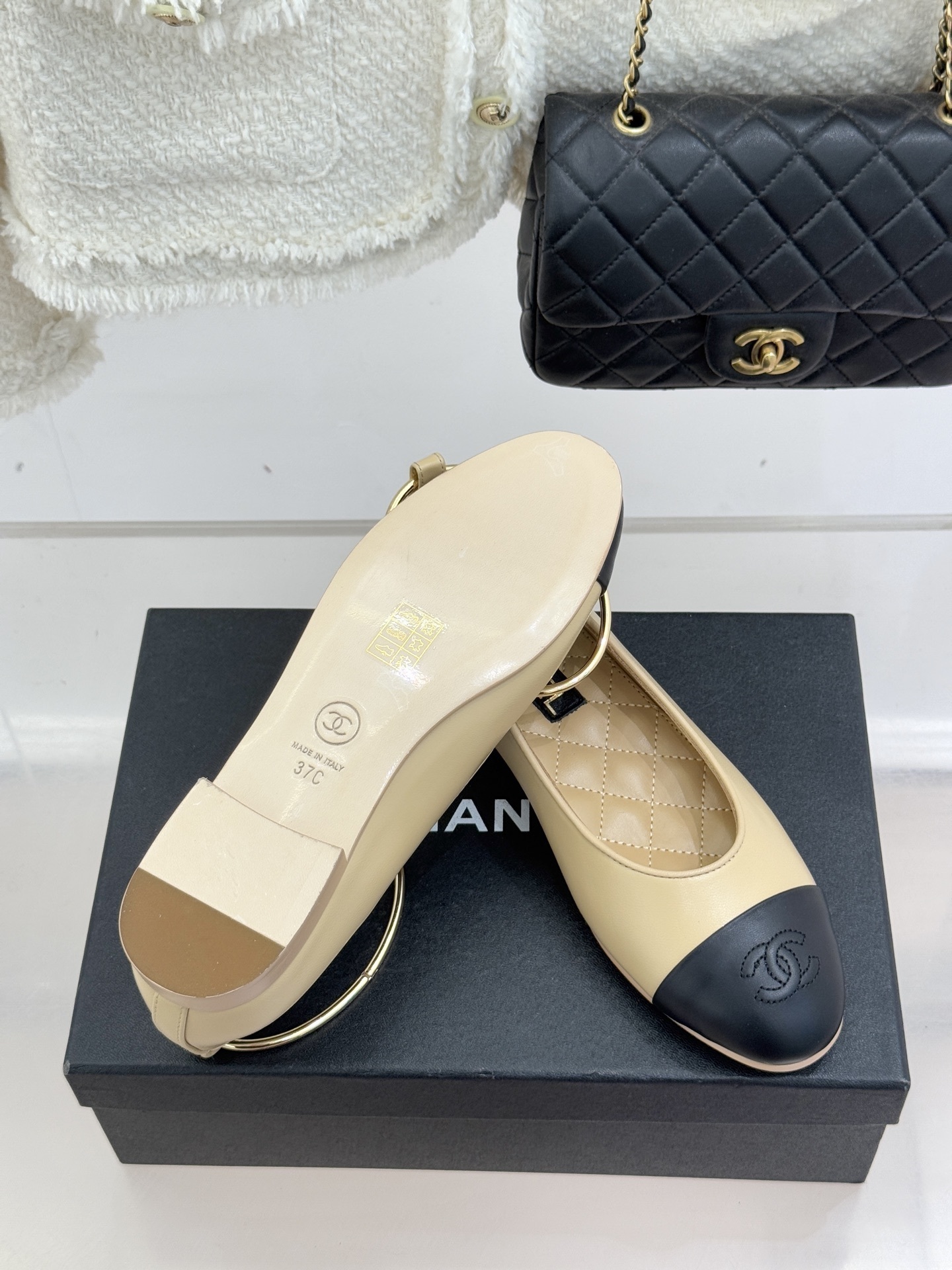 Pair of Chanel beige and black lambskin ballet flats with gold-tone ankle rings resting on a black box. - Hình 9