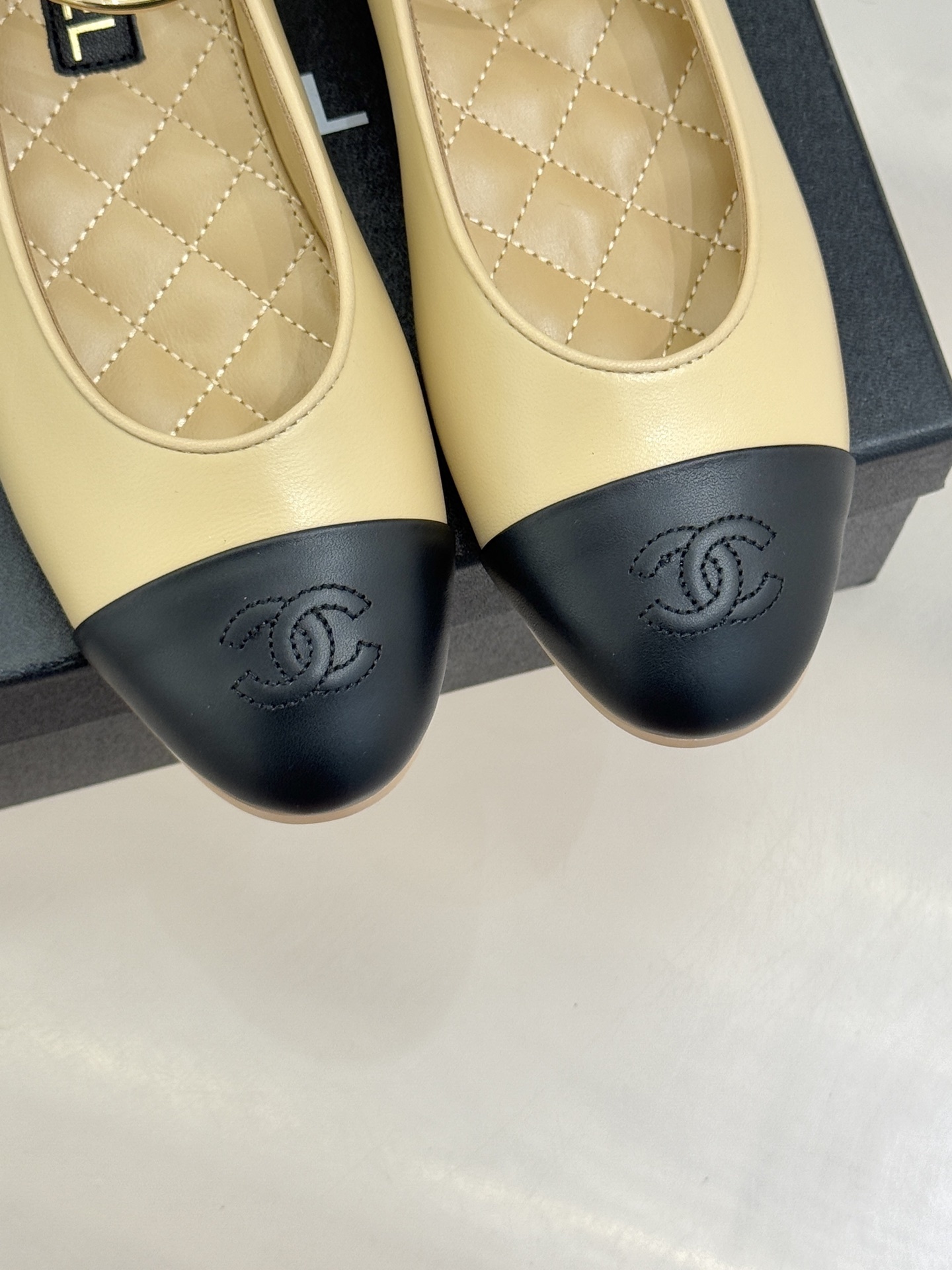 Pair of Chanel beige and black lambskin ballet flats with gold-tone ankle rings resting on a black box. - Hình 7