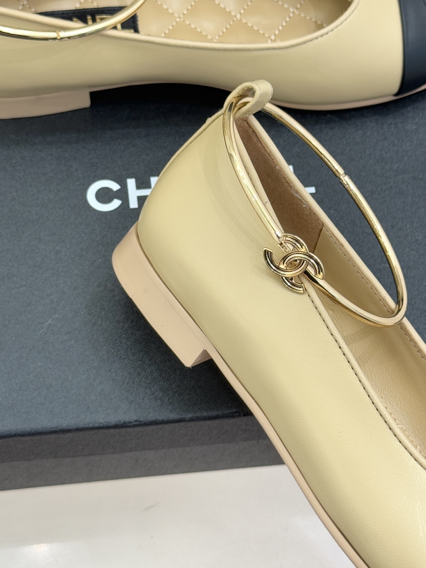 Pair of Chanel beige and black lambskin ballet flats with gold-tone ankle rings resting on a black box. - Hình 5
