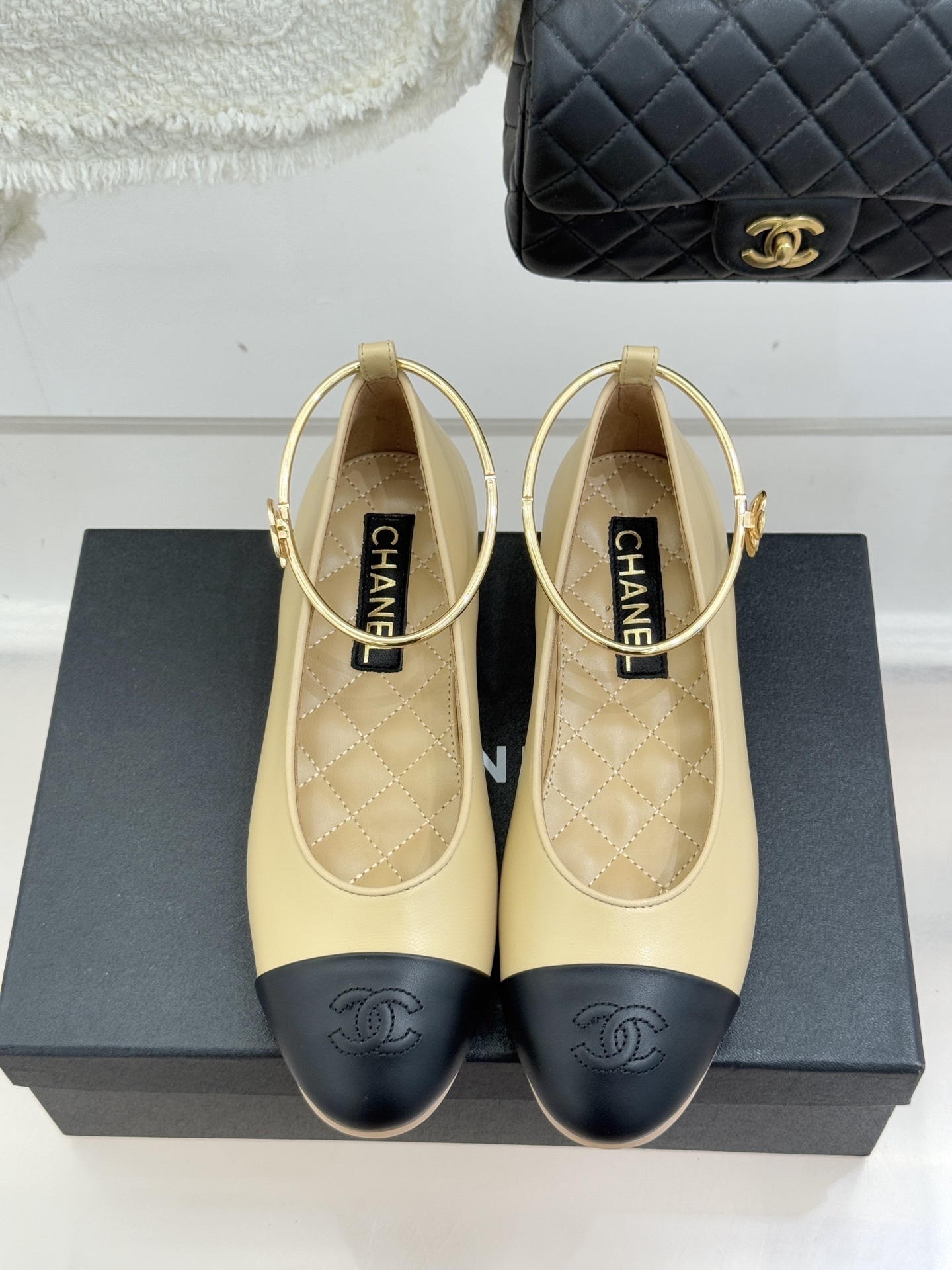 Pair of Chanel beige and black lambskin ballet flats with gold-tone ankle rings resting on a black box. - Hình 4