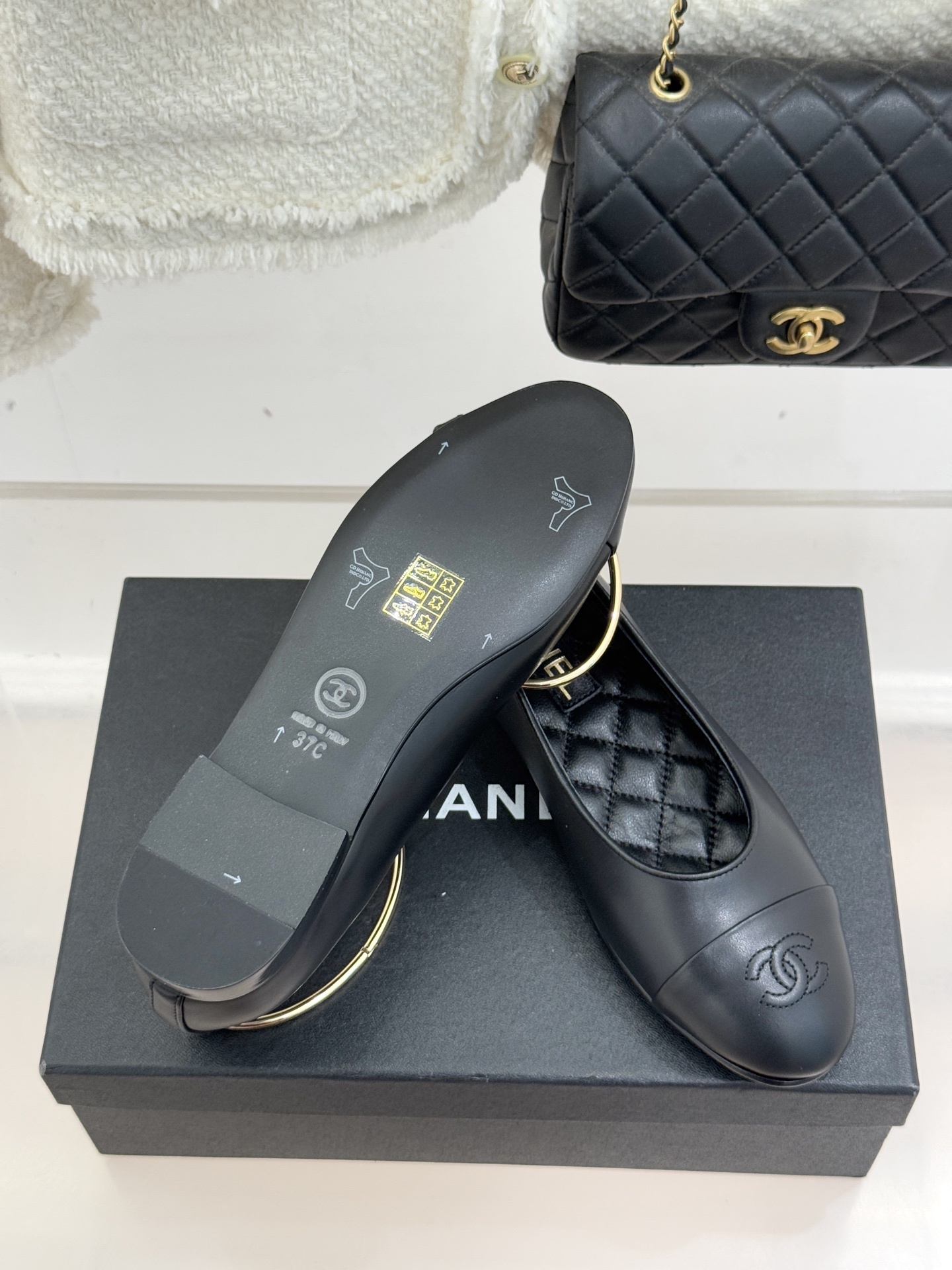A pair of luxury black leather ballet flats with gold ankle rings and embossed logos on a black designer box. - Hình 9