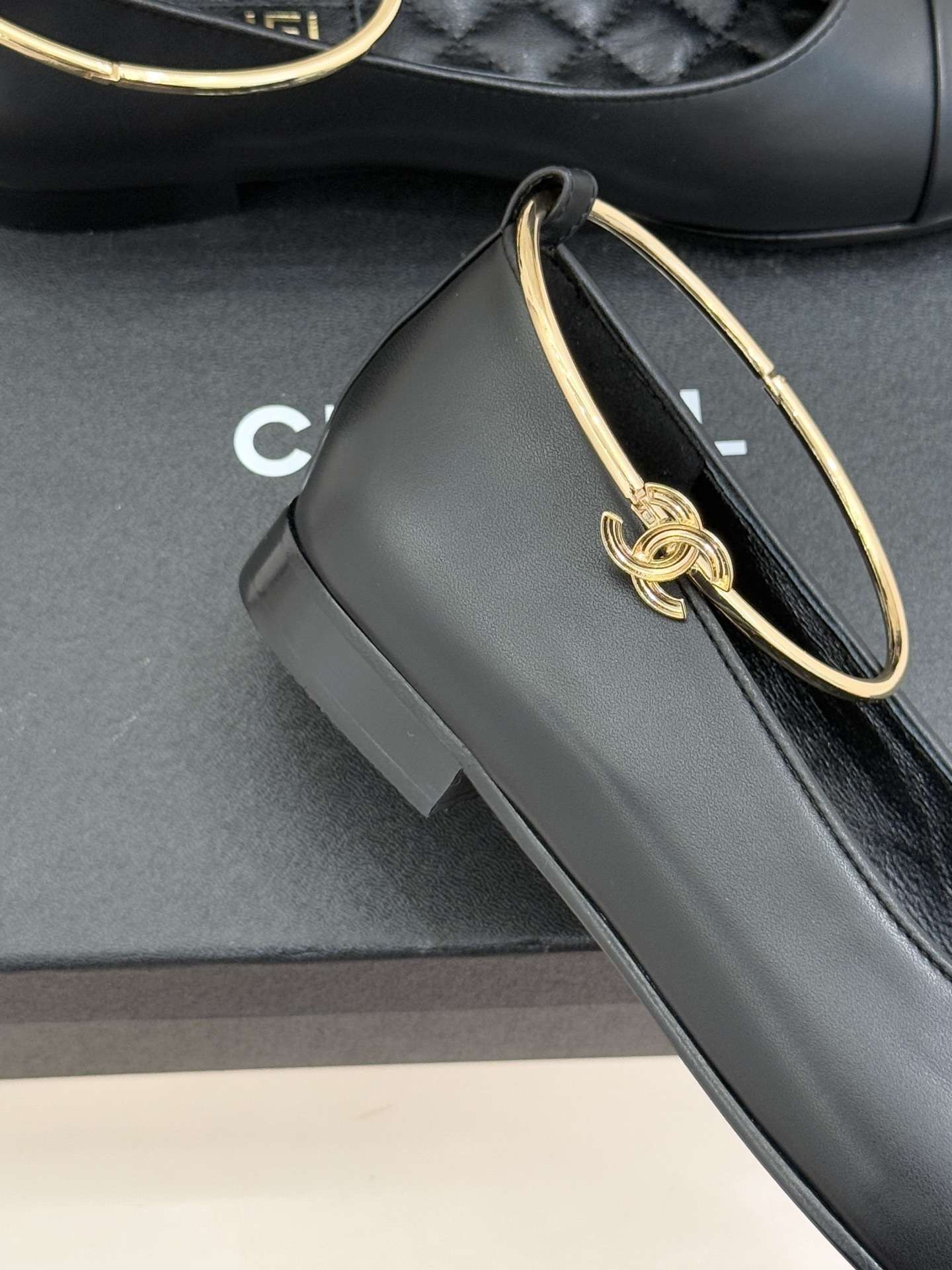 A pair of luxury black leather ballet flats with gold ankle rings and embossed logos on a black designer box. - Hình 6
