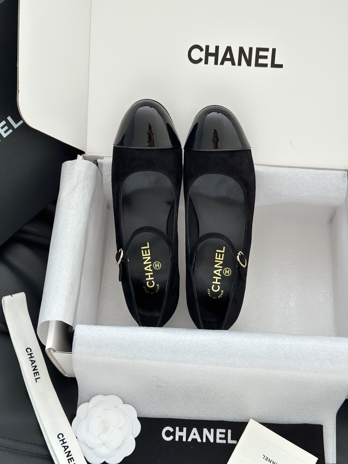 A pair of black Chanel suede Mary Jane pumps with patent leather cap toes and a distinctive white pearl heel displayed in a luxury white box. - Hình 6