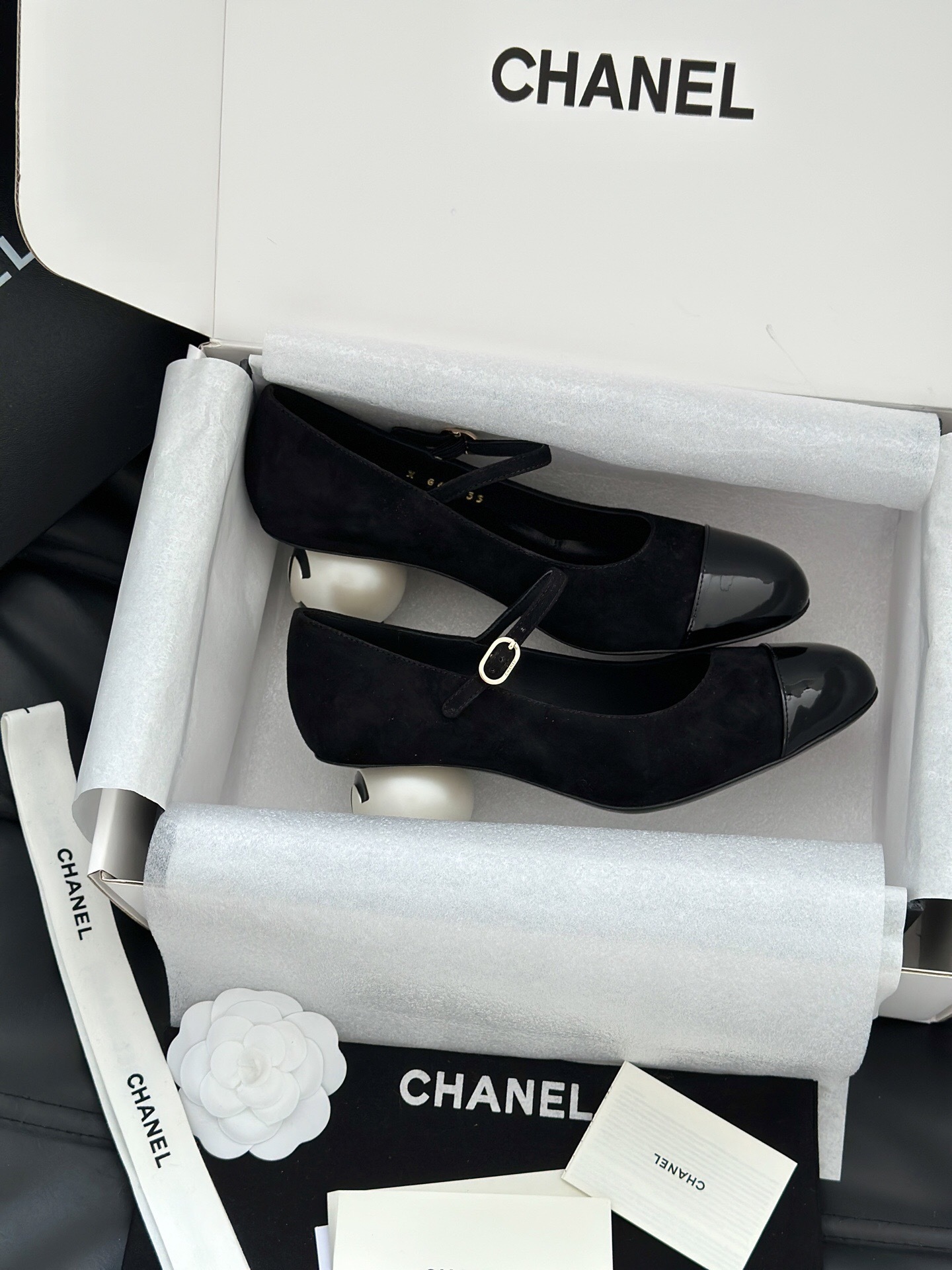 A pair of black Chanel suede Mary Jane pumps with patent leather cap toes and a distinctive white pearl heel displayed in a luxury white box. - Hình 5