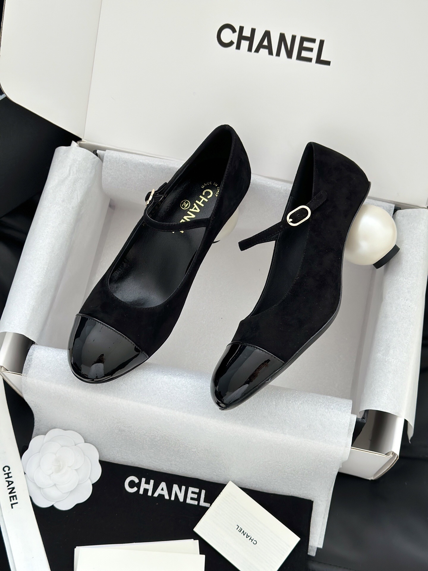 A pair of black Chanel suede Mary Jane pumps with patent leather cap toes and a distinctive white pearl heel displayed in a luxury white box. - Hình 4