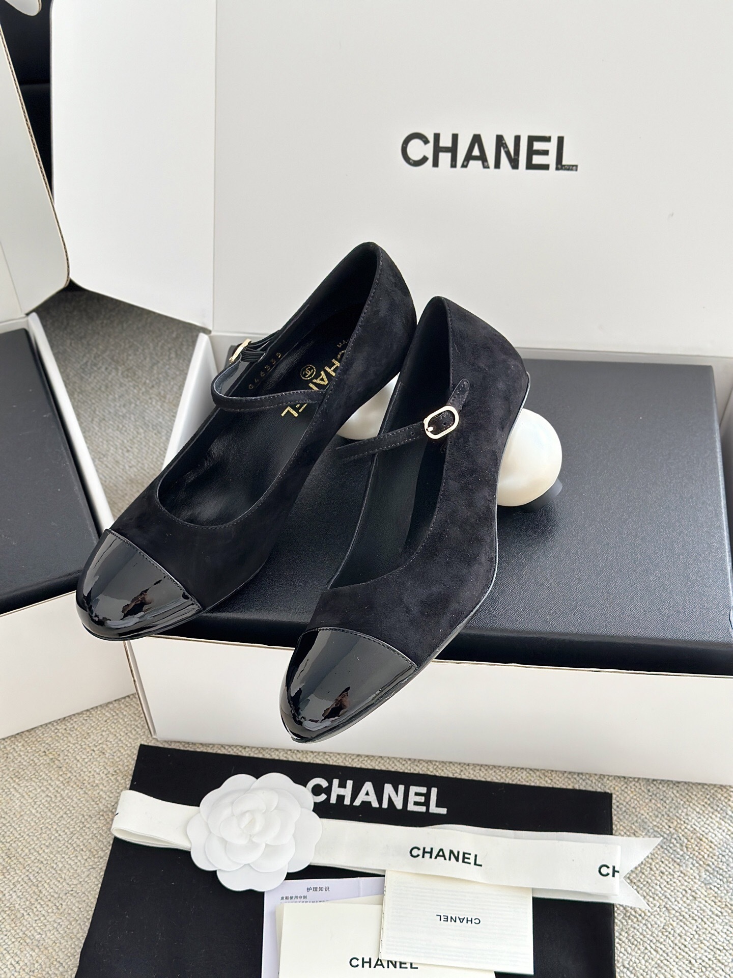 A pair of black Chanel suede Mary Jane pumps with patent leather cap toes and a distinctive white pearl heel displayed in a luxury white box. - Hình 3