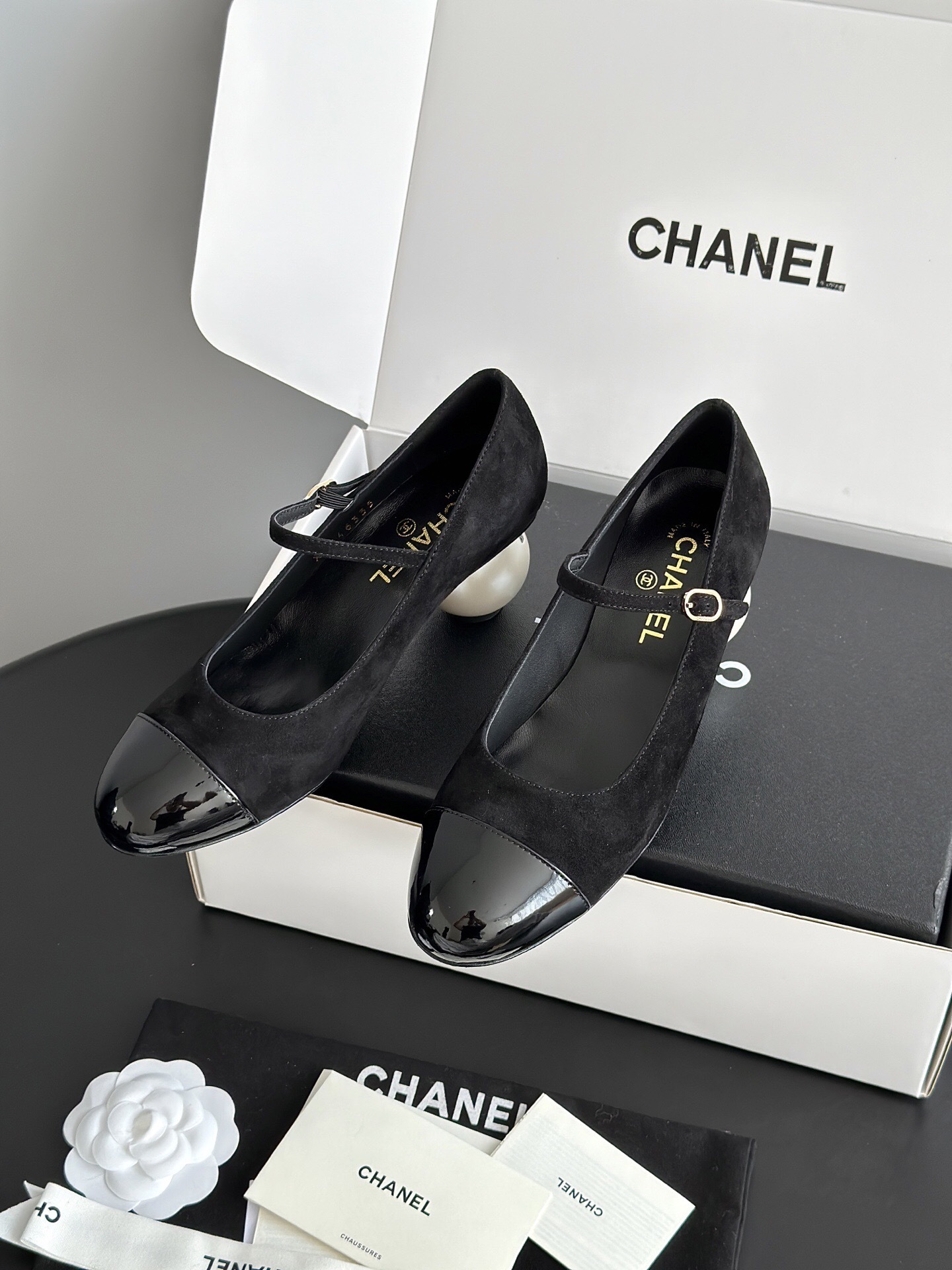 A pair of black Chanel suede Mary Jane pumps with patent leather cap toes and a distinctive white pearl heel displayed in a luxury white box. - Hình 2