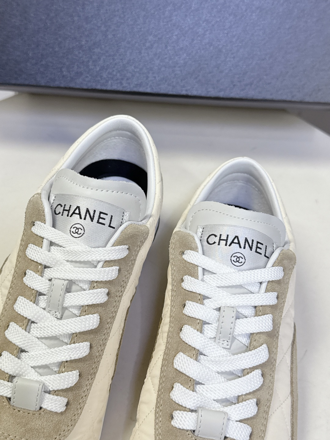 A pair of beige Chanel quilted low-top sneakers with suede accents and white laces next to a black Chanel shoe box. - Hình 9