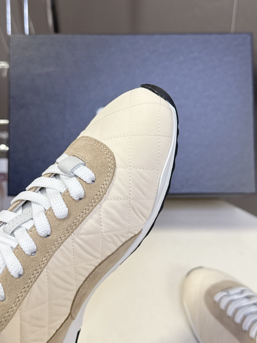 A pair of beige Chanel quilted low-top sneakers with suede accents and white laces next to a black Chanel shoe box. - Hình 7