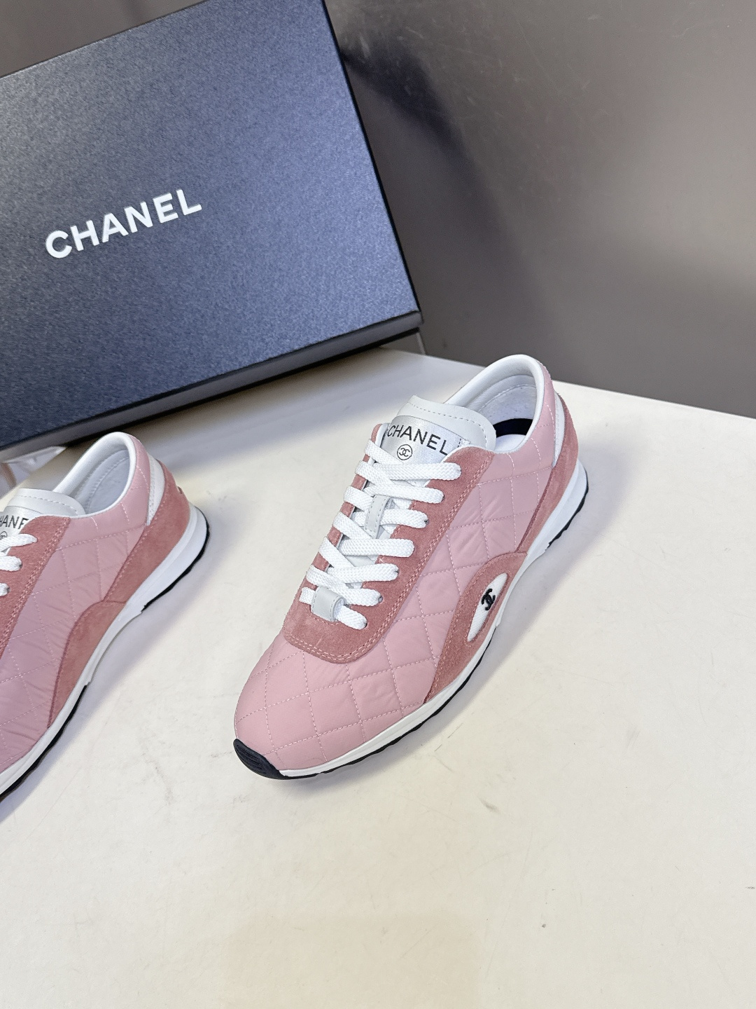 A pair of luxury Chanel pink quilted sneakers with suede trim and white laces placed next to a Chanel branded shoe box. - Hình 3