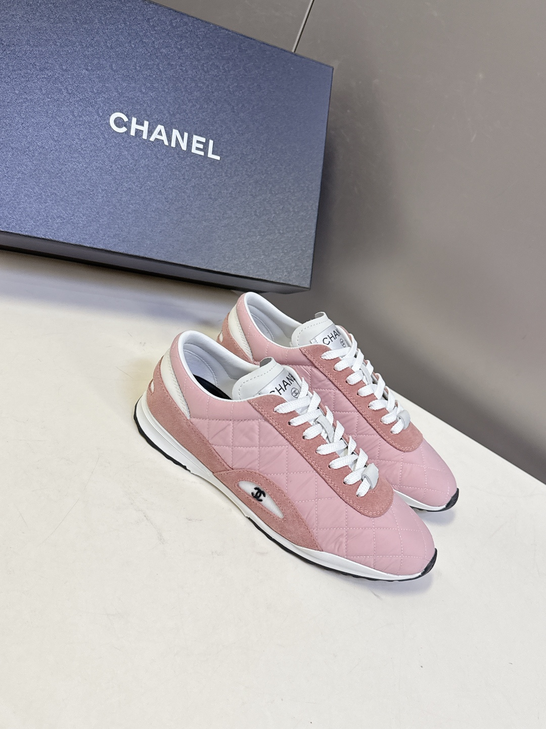 A pair of luxury Chanel pink quilted sneakers with suede trim and white laces placed next to a Chanel branded shoe box. - Hình 2