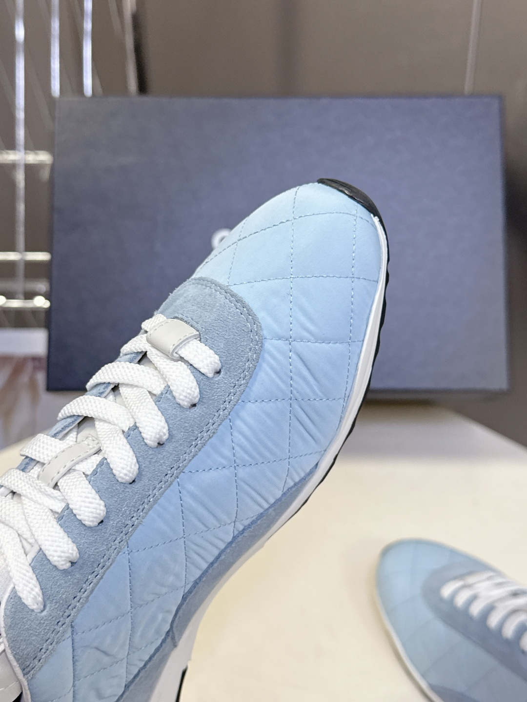 A pair of light blue quilted Chanel sneakers with suede details and white laces next to a black Chanel box - Hình 8