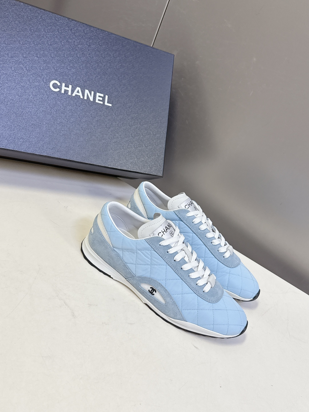 A pair of light blue quilted Chanel sneakers with suede details and white laces next to a black Chanel box - Hình 2