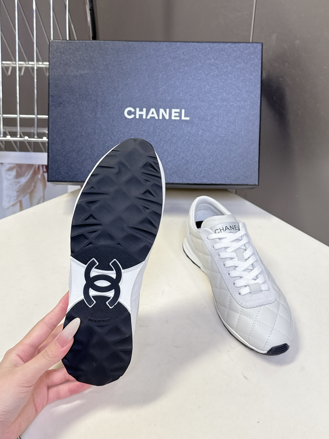 A pair of white Chanel quilted leather sneakers with suede panels and CC logo shown next to their original black Chanel box. - Hình 9
