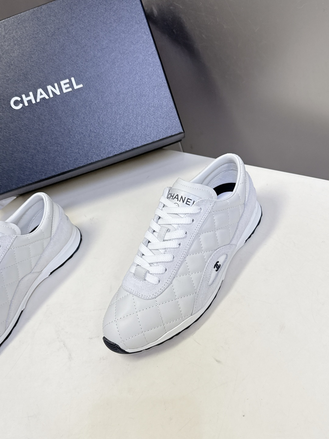 A pair of white Chanel quilted leather sneakers with suede panels and CC logo shown next to their original black Chanel box. - Hình 3
