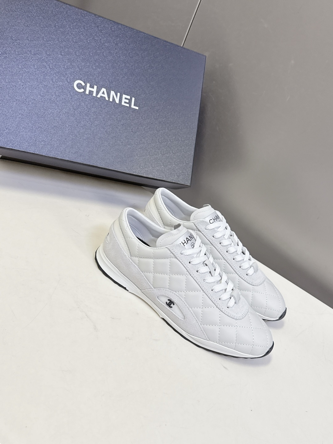 A pair of white Chanel quilted leather sneakers with suede panels and CC logo shown next to their original black Chanel box. - Hình 2