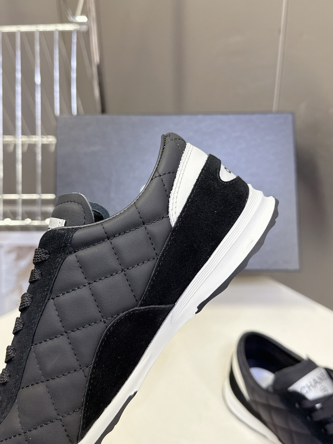 A pair of black Chanel quilted sneakers with white accents and CC logo next to a black Chanel shoe box. - Hình 8