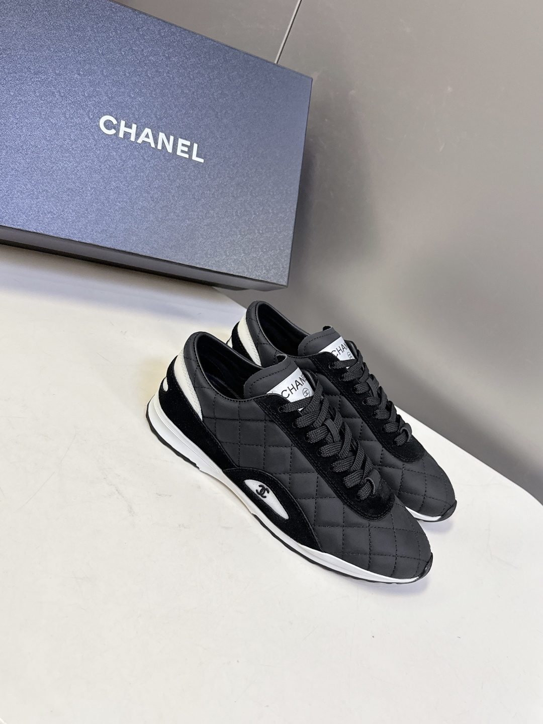 A pair of black Chanel quilted sneakers with white accents and CC logo next to a black Chanel shoe box. - Hình 2