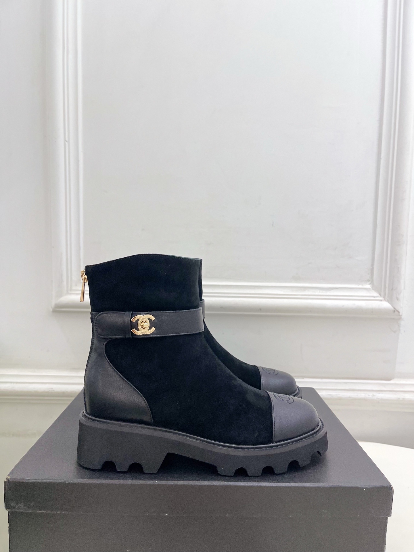 Pair of black Chanel suede ankle boots with leather toe caps and gold CC logo buckles resting on a black box. - Hình 2