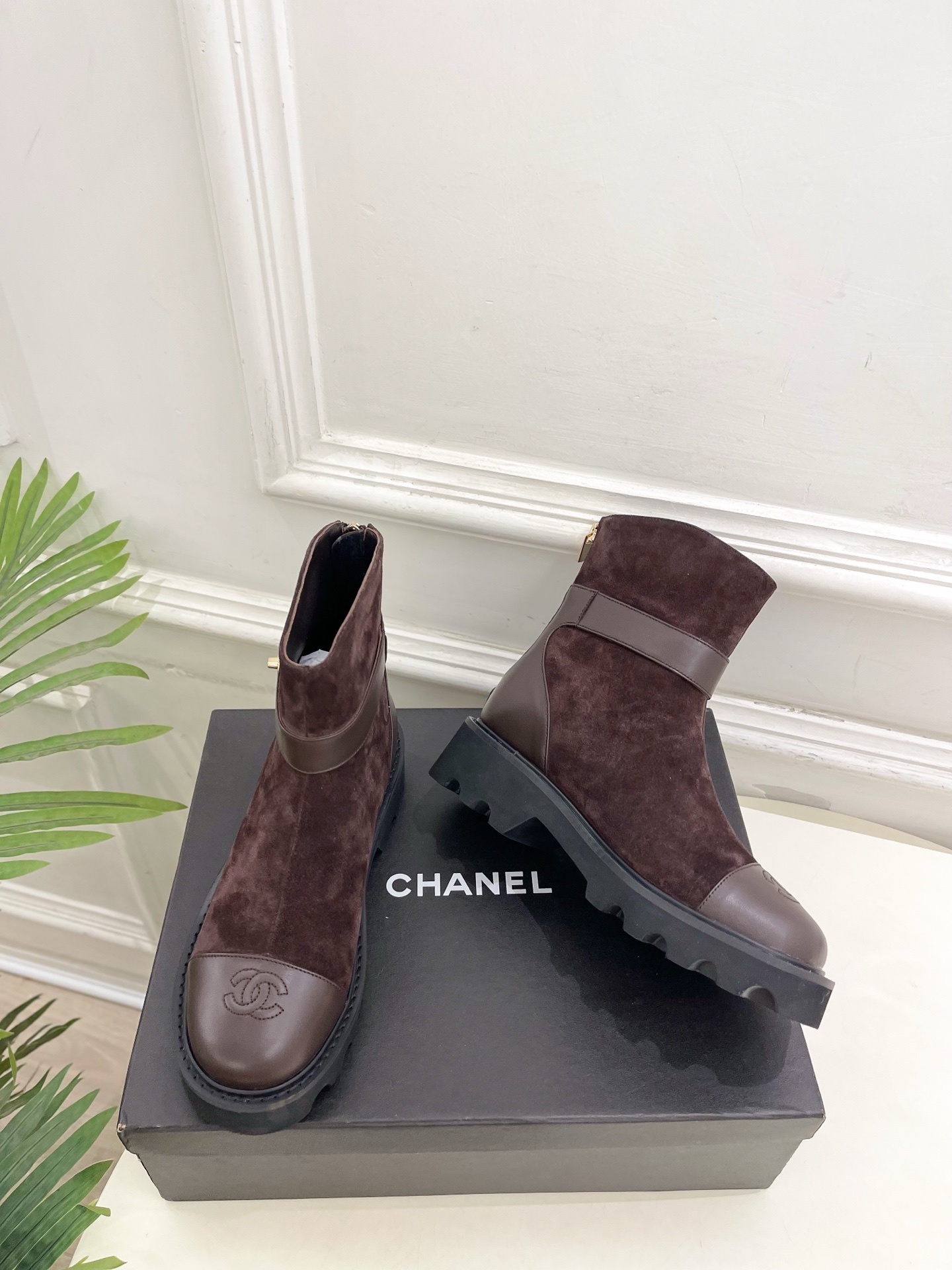 Pair of brown suede Chanel ankle boots with gold CC logo hardware and chunky black lug soles placed on a black shoe box. - Hình 5