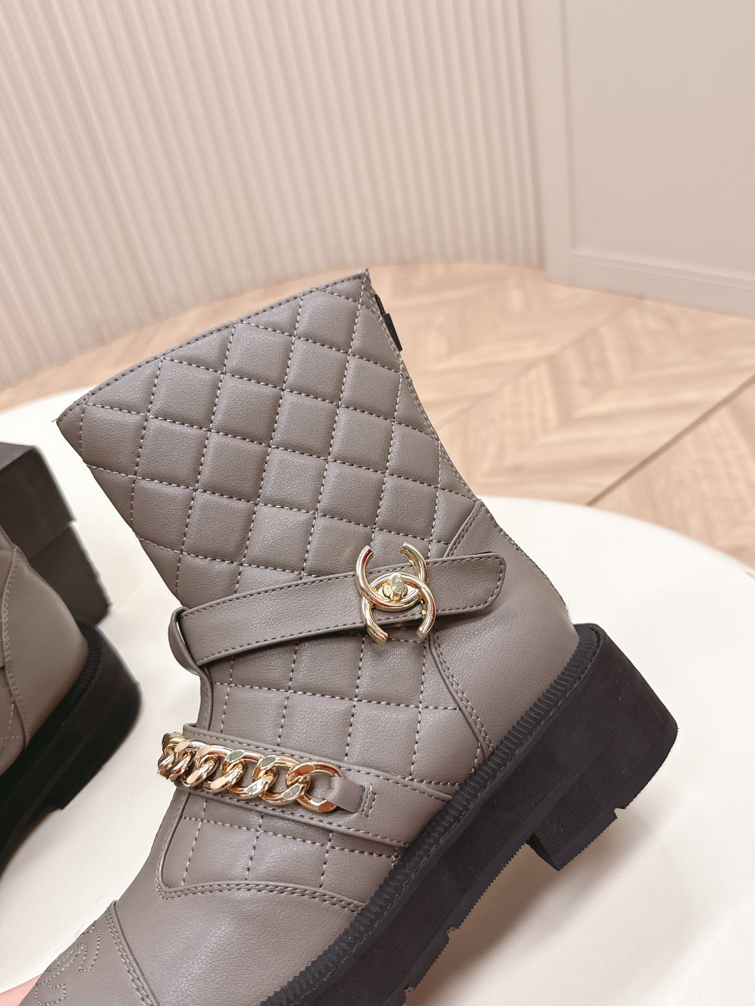 A pair of grey Chanel quilted leather boots with gold chain details and CC logo displayed on a white surface next to a Chanel shoe box. - Hình 8