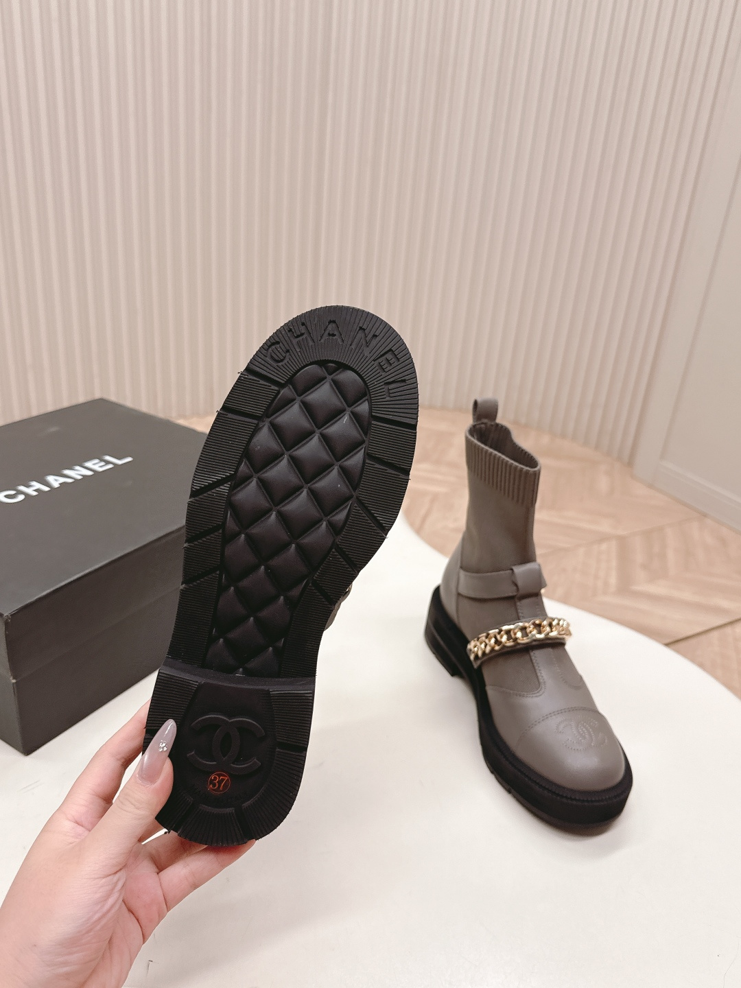 A pair of grey Chanel quilted leather boots with gold chain details and CC logo displayed on a white surface next to a Chanel shoe box. - Hình 6