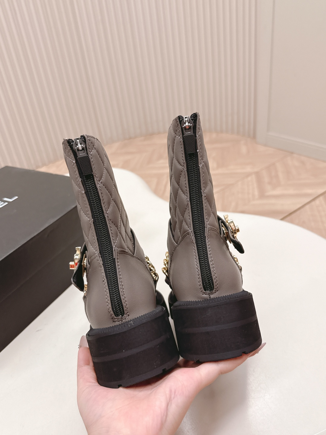 A pair of grey Chanel quilted leather boots with gold chain details and CC logo displayed on a white surface next to a Chanel shoe box. - Hình 5