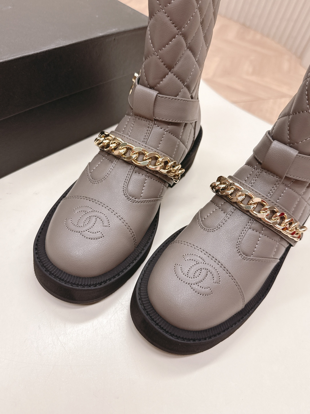 A pair of grey Chanel quilted leather boots with gold chain details and CC logo displayed on a white surface next to a Chanel shoe box. - Hình 4