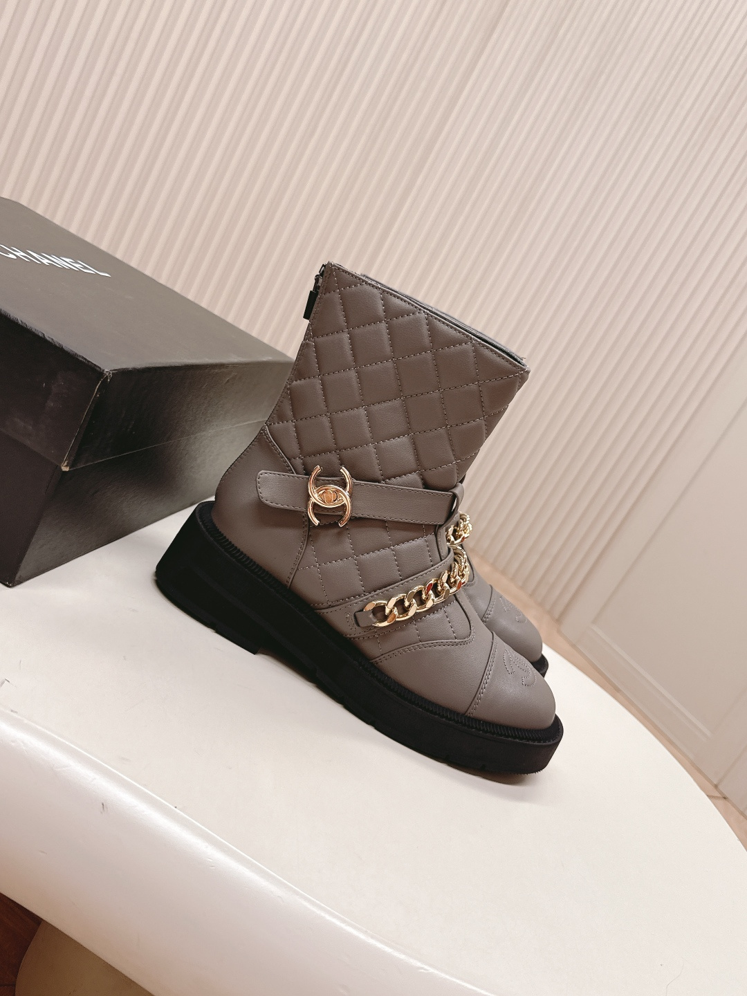 A pair of grey Chanel quilted leather boots with gold chain details and CC logo displayed on a white surface next to a Chanel shoe box. - Hình 2