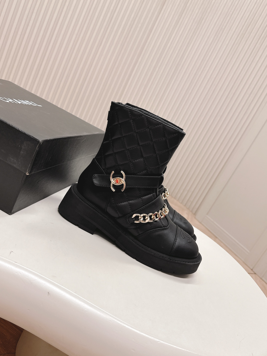 A pair of Chanel black quilted leather ankle boots with gold chain and buckle detail placed on a white surface next to a black shoe box. - Hình 8