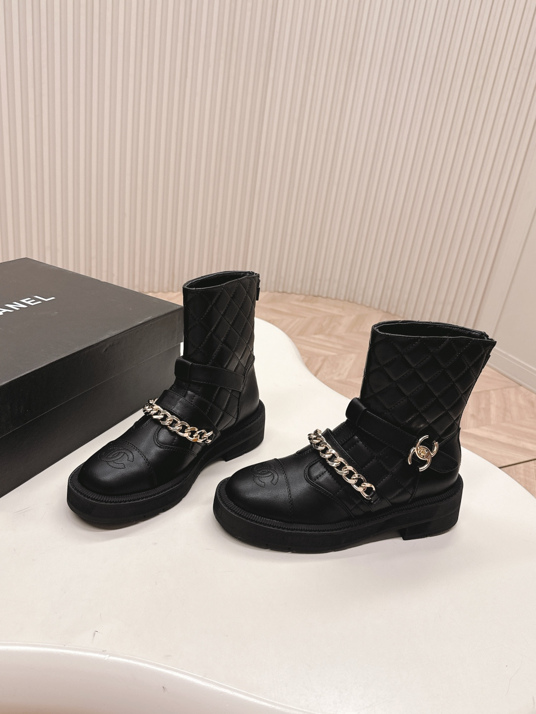 A pair of Chanel black quilted leather ankle boots with gold chain and buckle detail placed on a white surface next to a black shoe box. - Hình 7