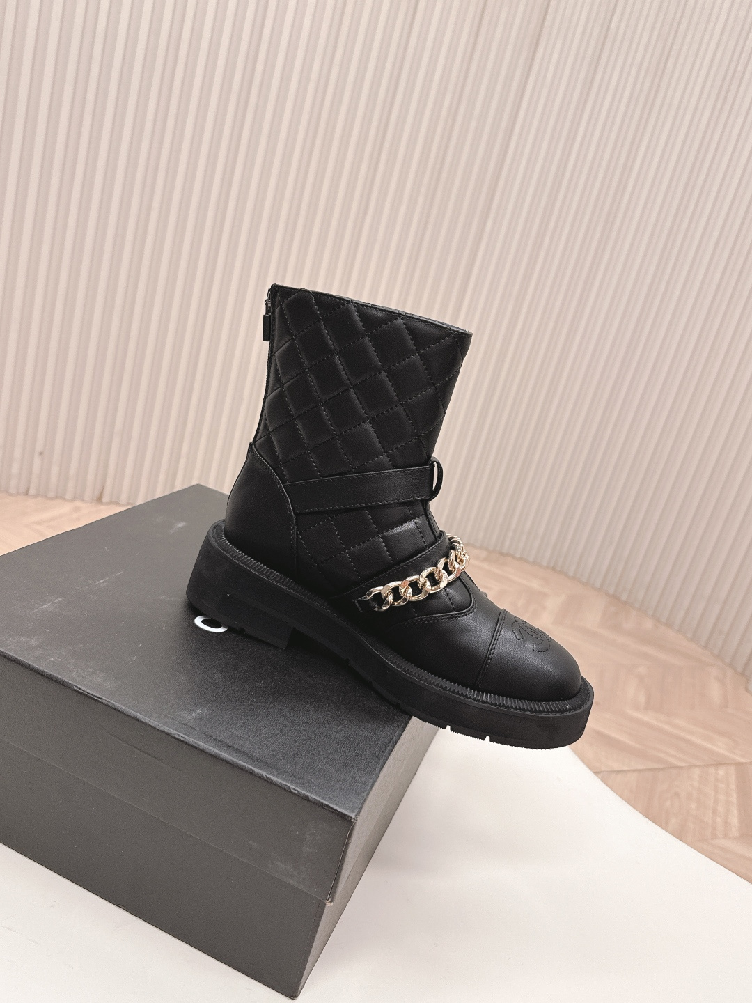 A pair of Chanel black quilted leather ankle boots with gold chain and buckle detail placed on a white surface next to a black shoe box. - Hình 6