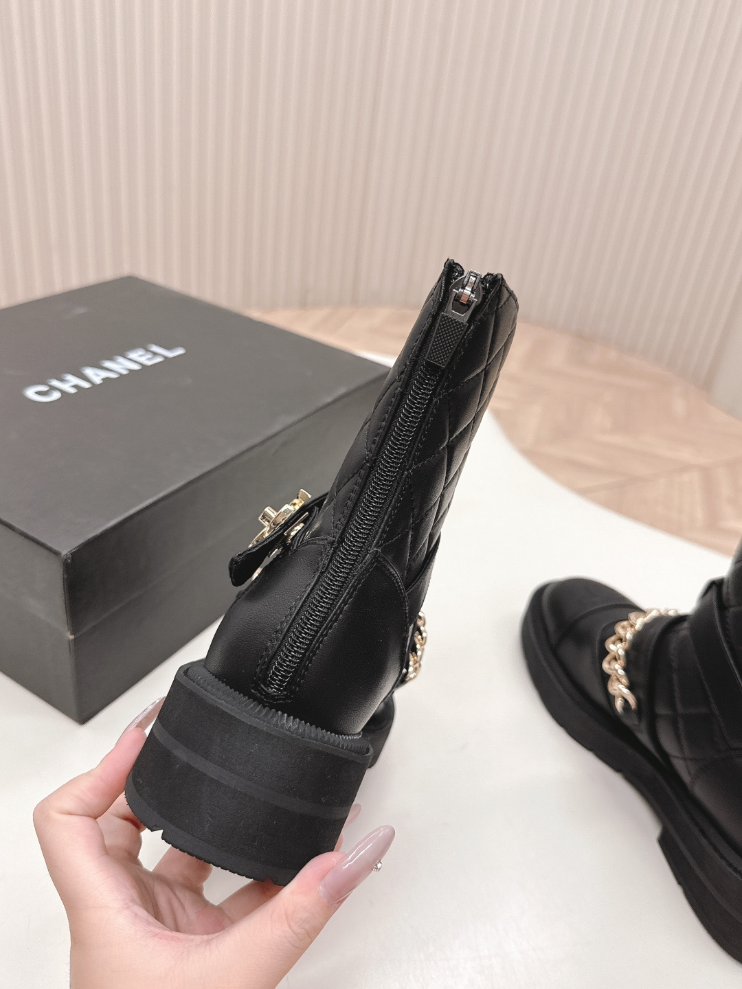 A pair of Chanel black quilted leather ankle boots with gold chain and buckle detail placed on a white surface next to a black shoe box. - Hình 4
