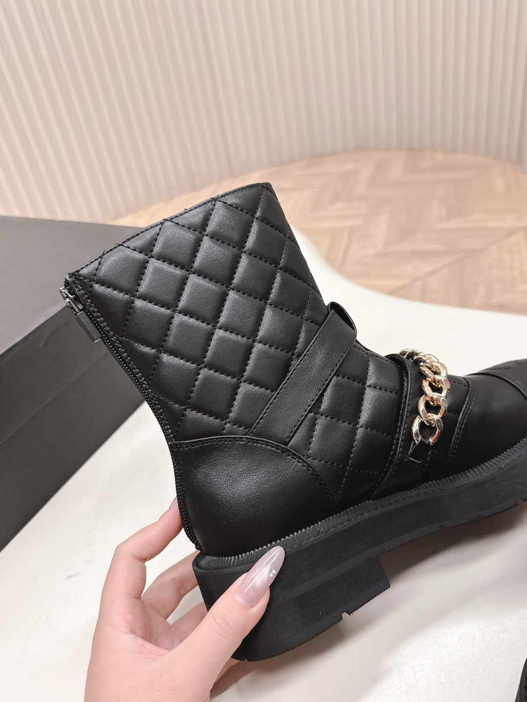 A pair of Chanel black quilted leather ankle boots with gold chain and buckle detail placed on a white surface next to a black shoe box. - Hình 3