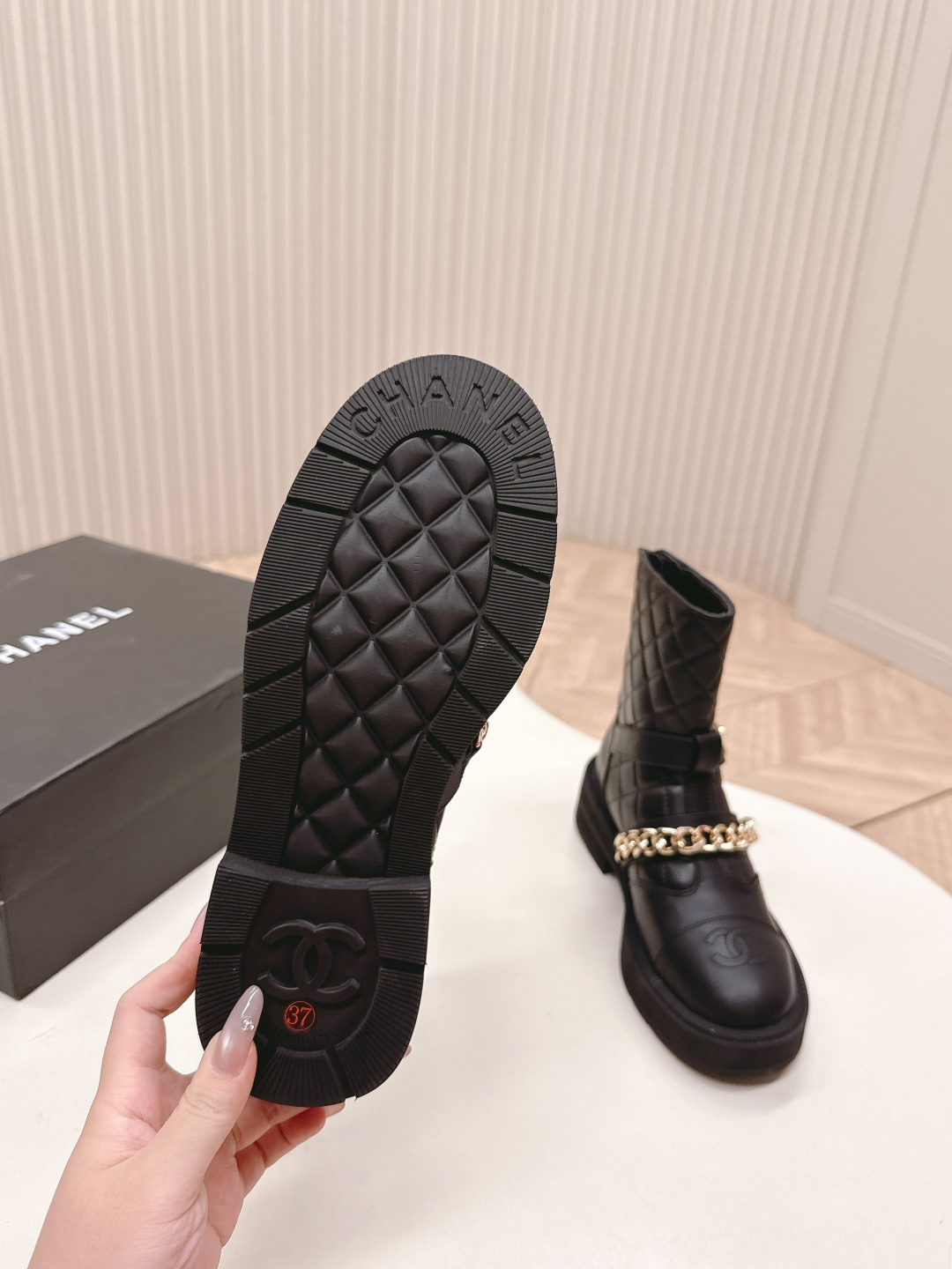 A pair of Chanel black quilted leather ankle boots with gold chain and buckle detail placed on a white surface next to a black shoe box. - Hình 2
