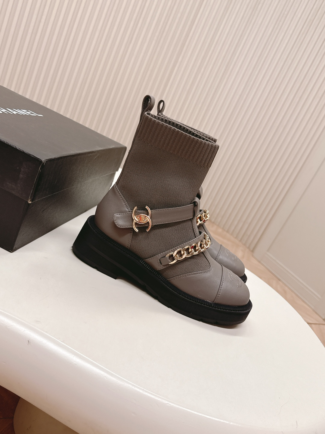Pair of grey Chanel sock boots with gold chain details and CC logo on a white table near a Chanel box. - Hình 4