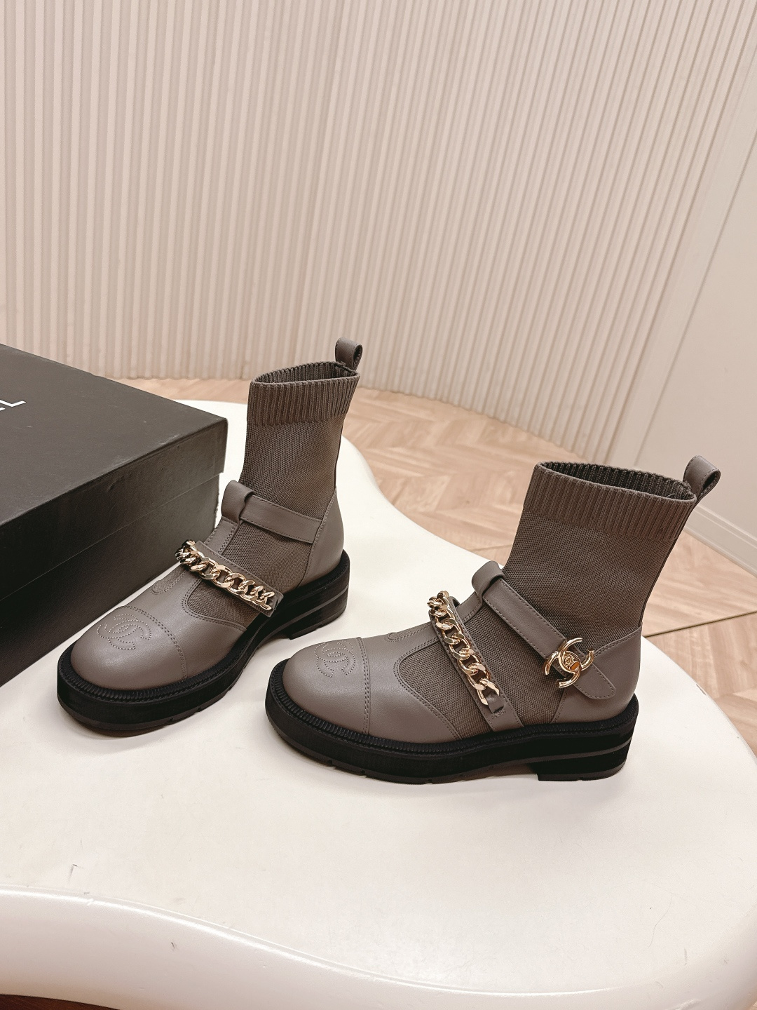 Pair of grey Chanel sock boots with gold chain details and CC logo on a white table near a Chanel box. - Hình 3