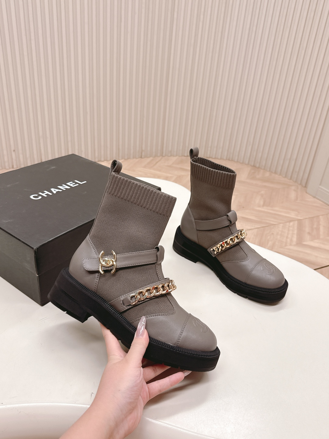 Pair of grey Chanel sock boots with gold chain details and CC logo on a white table near a Chanel box. - Hình 2
